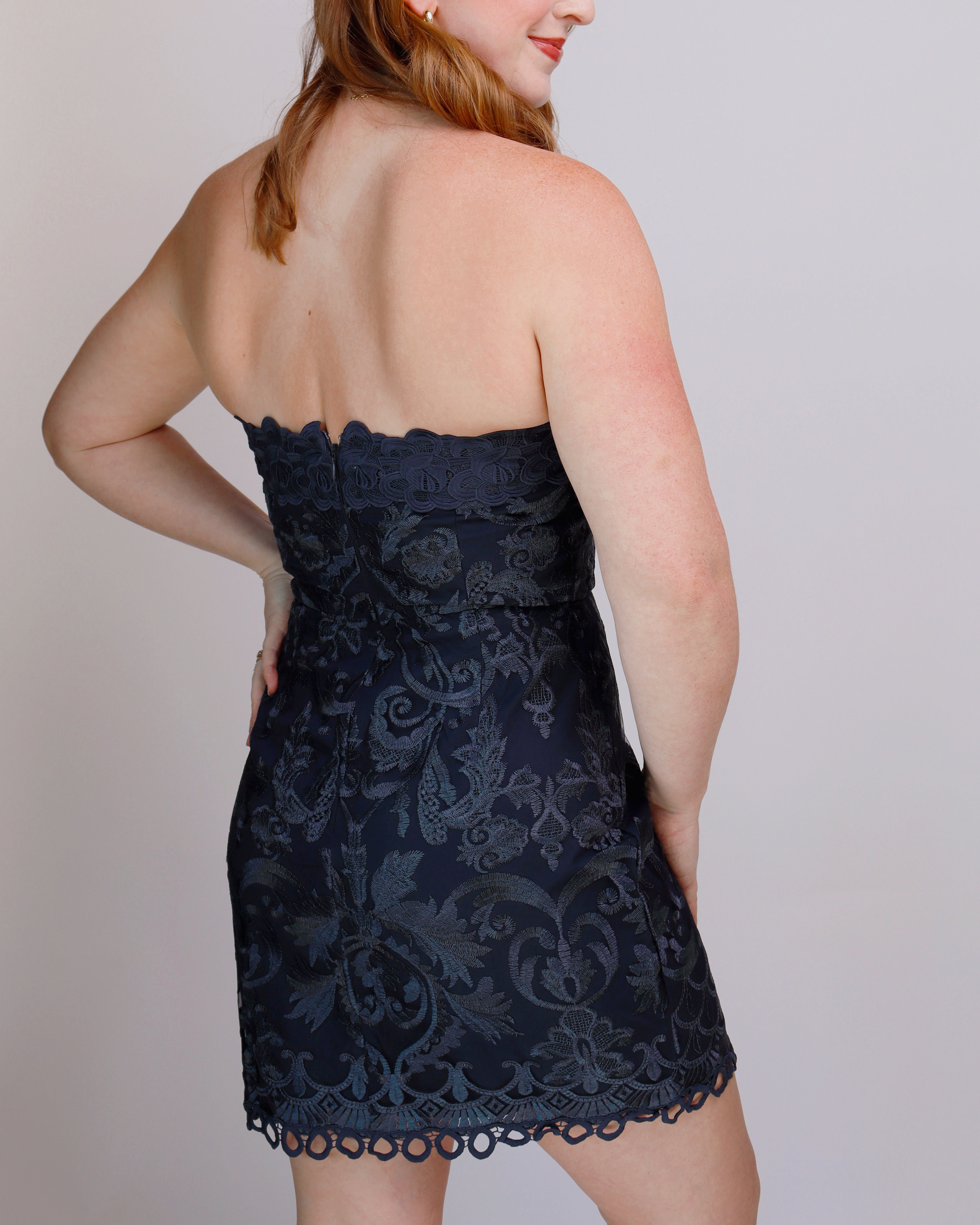 A woman wearing a strapless lace mini dress, shown from the back. The dress is dark navy blue with a tonal floral lace pattern and scalloped edges, photographed against a plain light gray background.