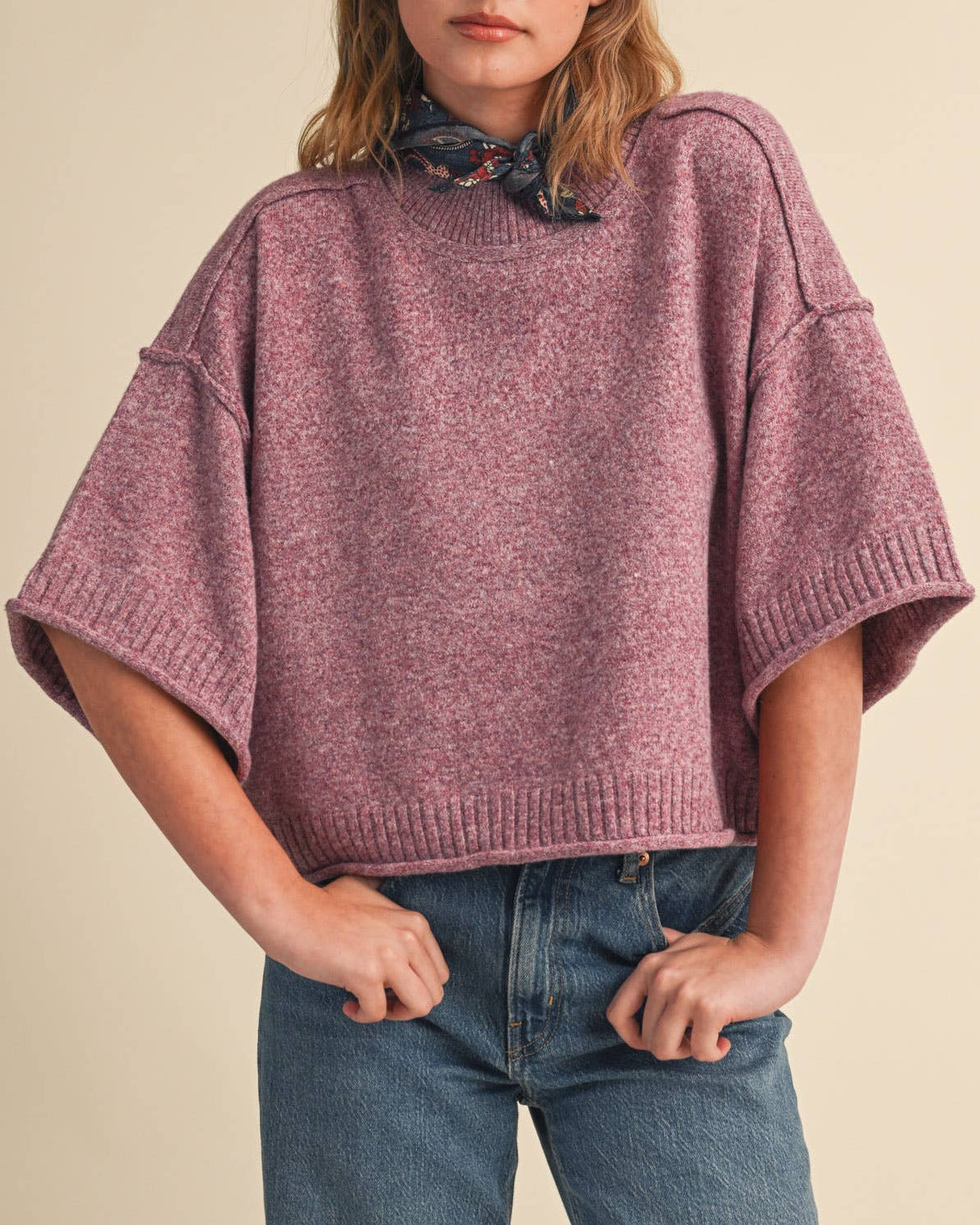 A woman wearing a heathered dusty rose knit sweater with wide three-quarter sleeves and a patterned neck scarf, paired with blue jeans, against a solid beige background.