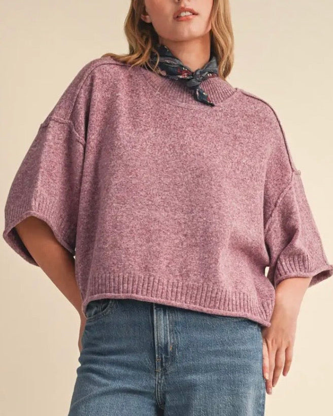 A woman wearing a heathered dusty rose knit sweater with wide three-quarter sleeves and a draped collar, paired with blue jeans and a dark patterned neck scarf, against a solid beige background.