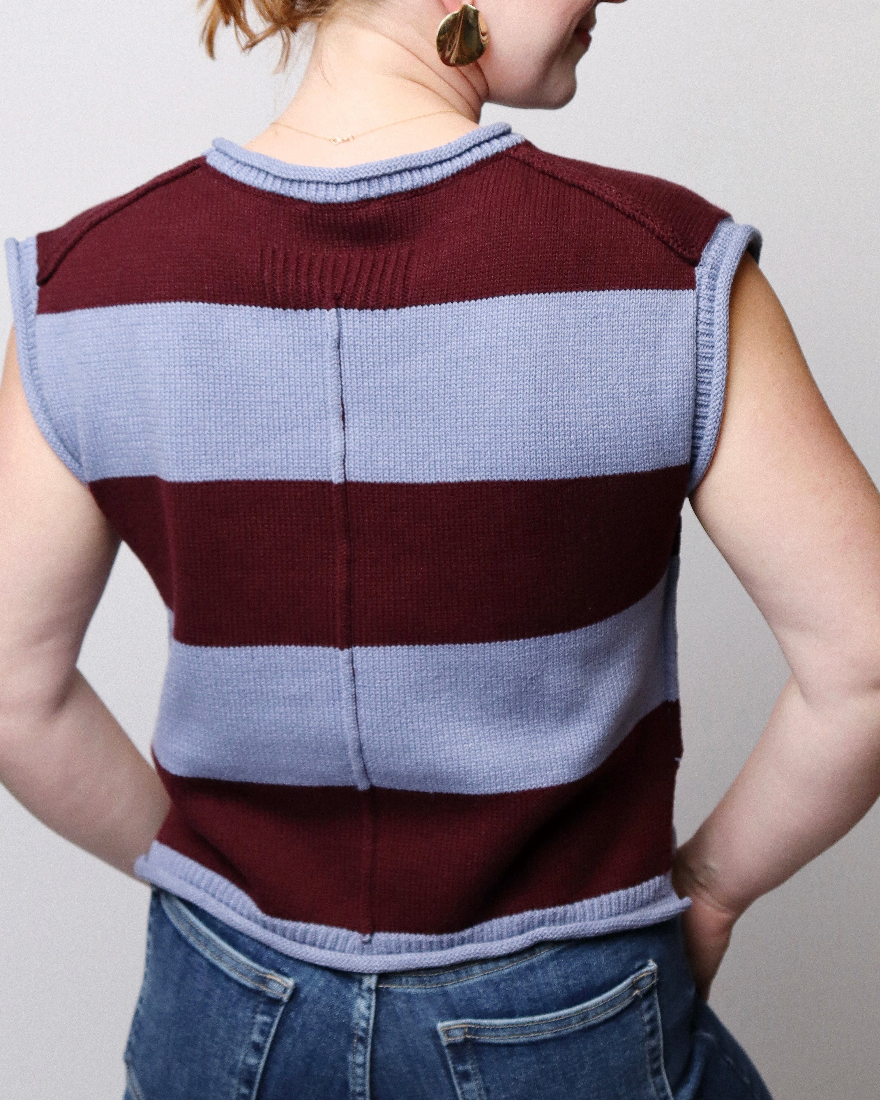 A person shown from the back wearing a sleeveless ribbed knit sweater vest with horizontal burgundy and light blue stripes, paired with blue jeans against a plain light gray background.