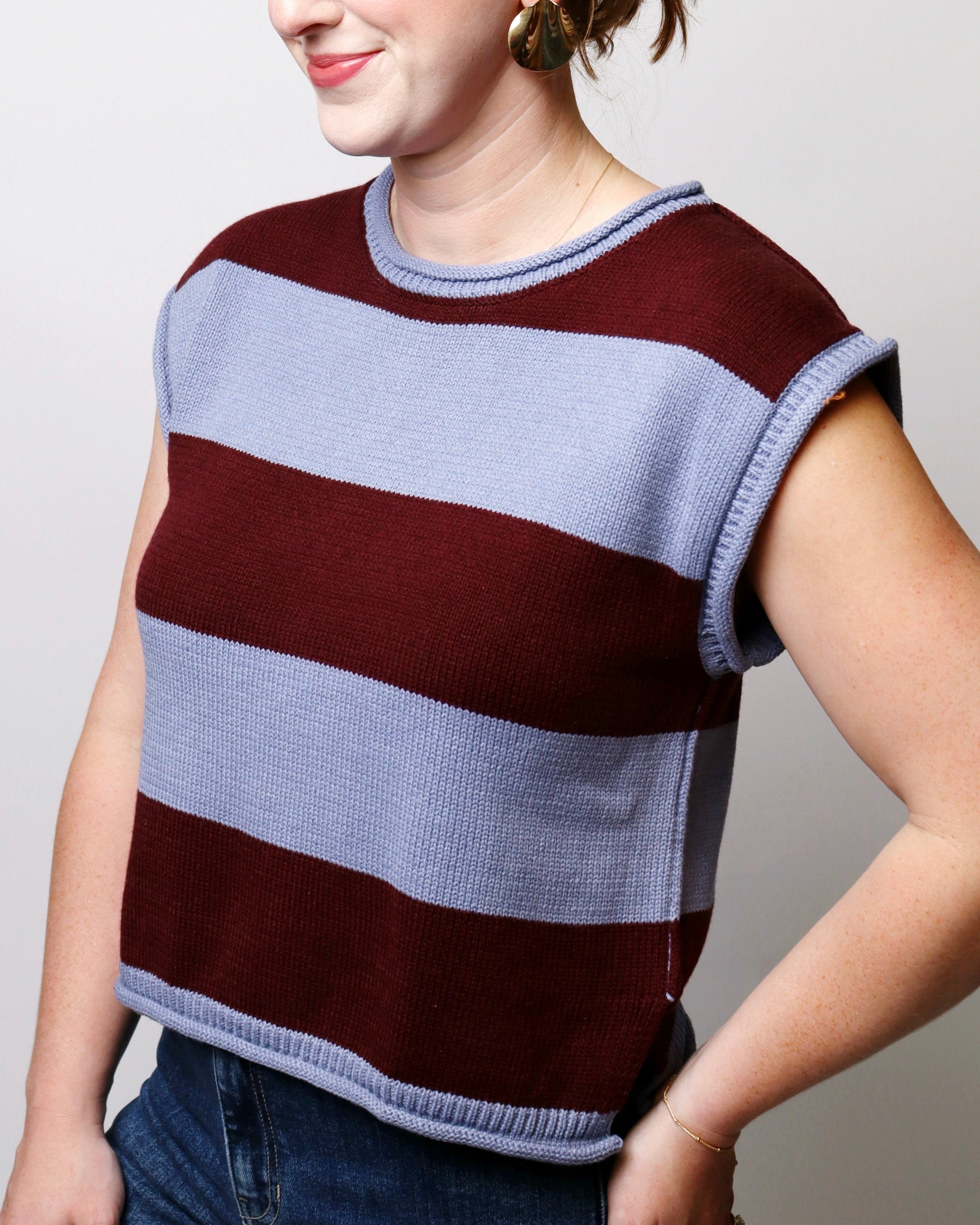 A woman wearing a sleeveless ribbed knit sweater with wide horizontal stripes in burgundy and light periwinkle blue, paired with blue jeans against a plain light gray background.
