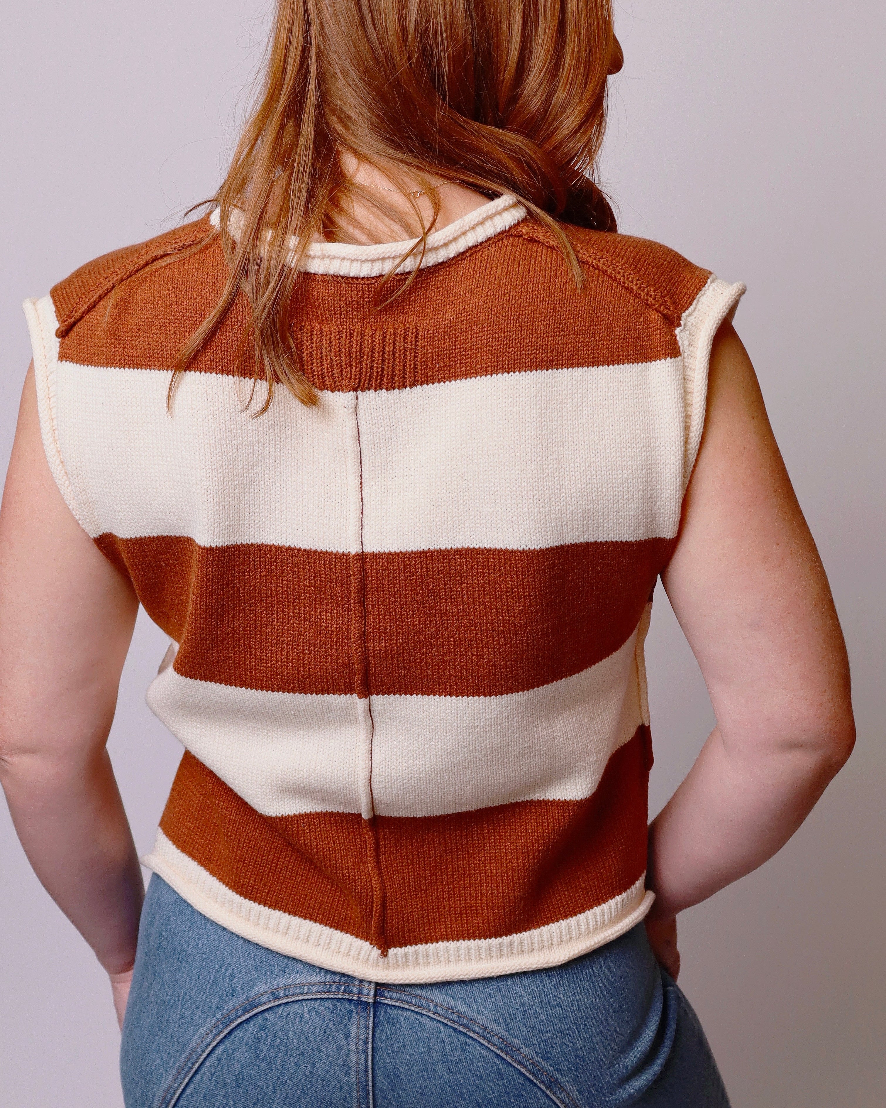 A woman viewed from the back wearing a sleeveless ribbed knit sweater vest with wide horizontal rust-brown and cream stripes, paired with blue jeans against a plain light gray background.