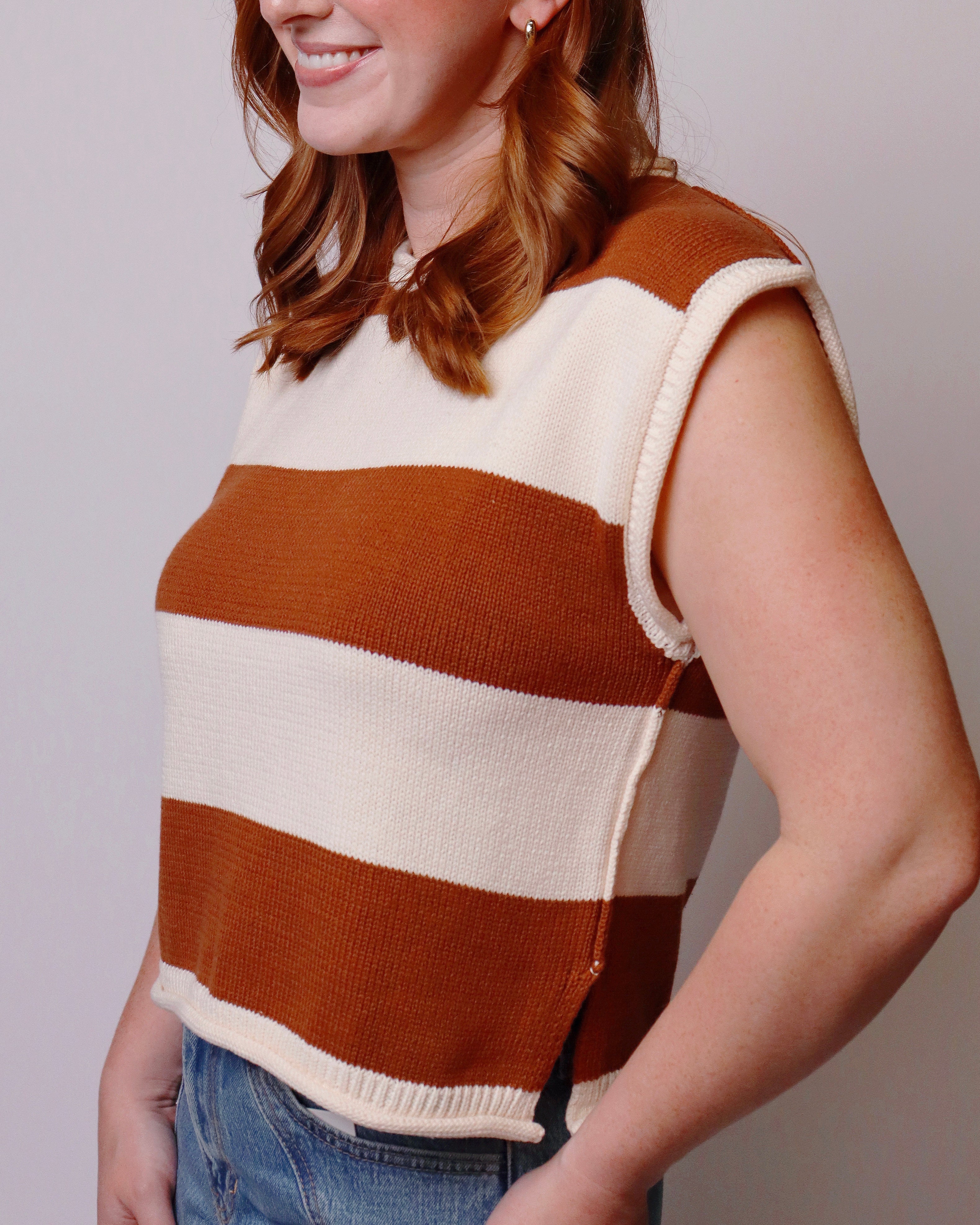 A woman wearing a sleeveless knit sweater vest with horizontal stripes in rust brown and cream, paired with blue jeans against a plain light gray background.