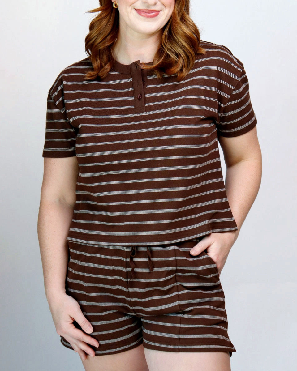 A woman wearing a matching brown and gray striped lounge set consisting of a short-sleeve henley top and drawstring shorts, photographed against a light gray studio background.