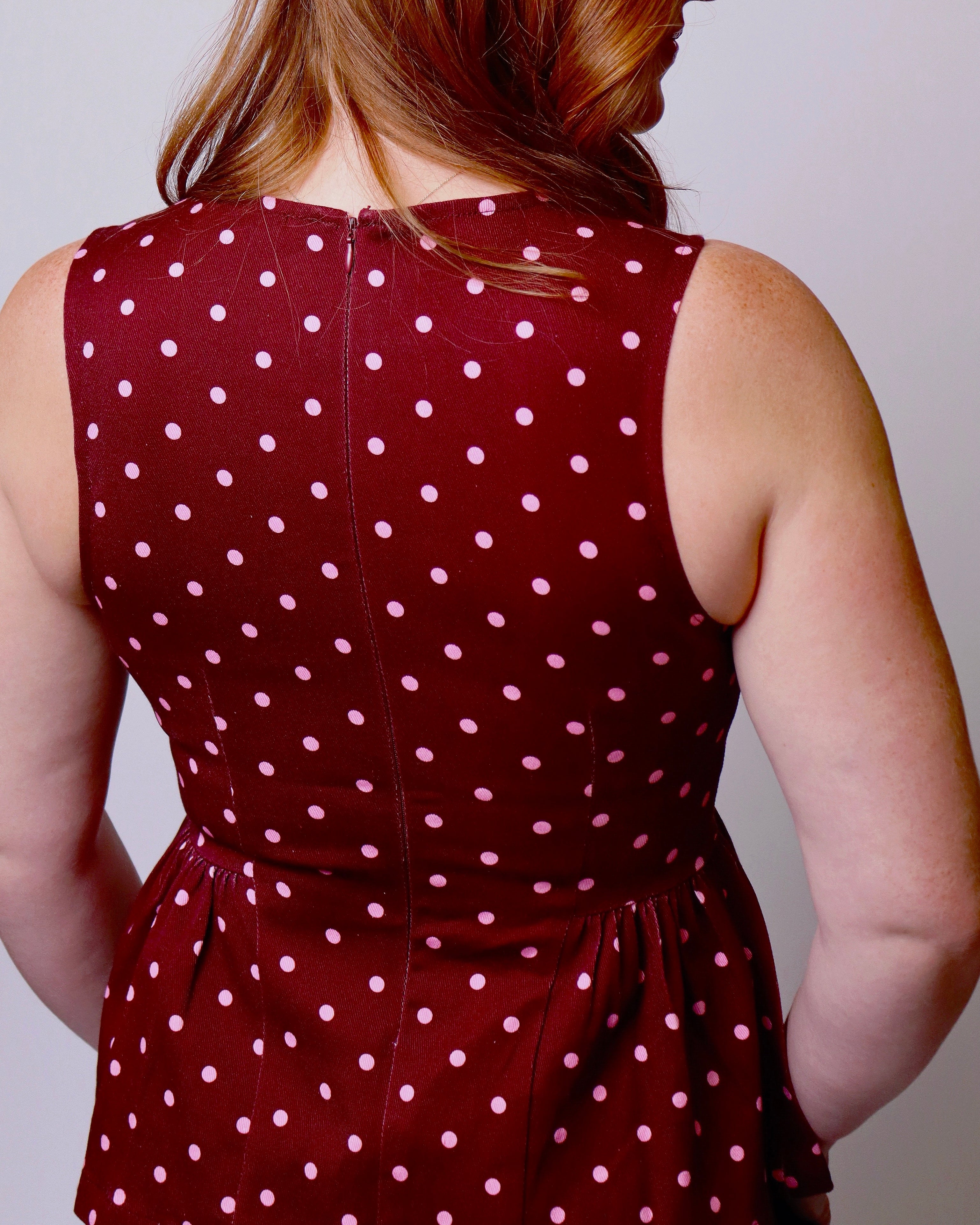 A woman viewed from the back wearing a sleeveless burgundy dress with a pink polka dot pattern and a matching waist tie, set against a plain light gray background.