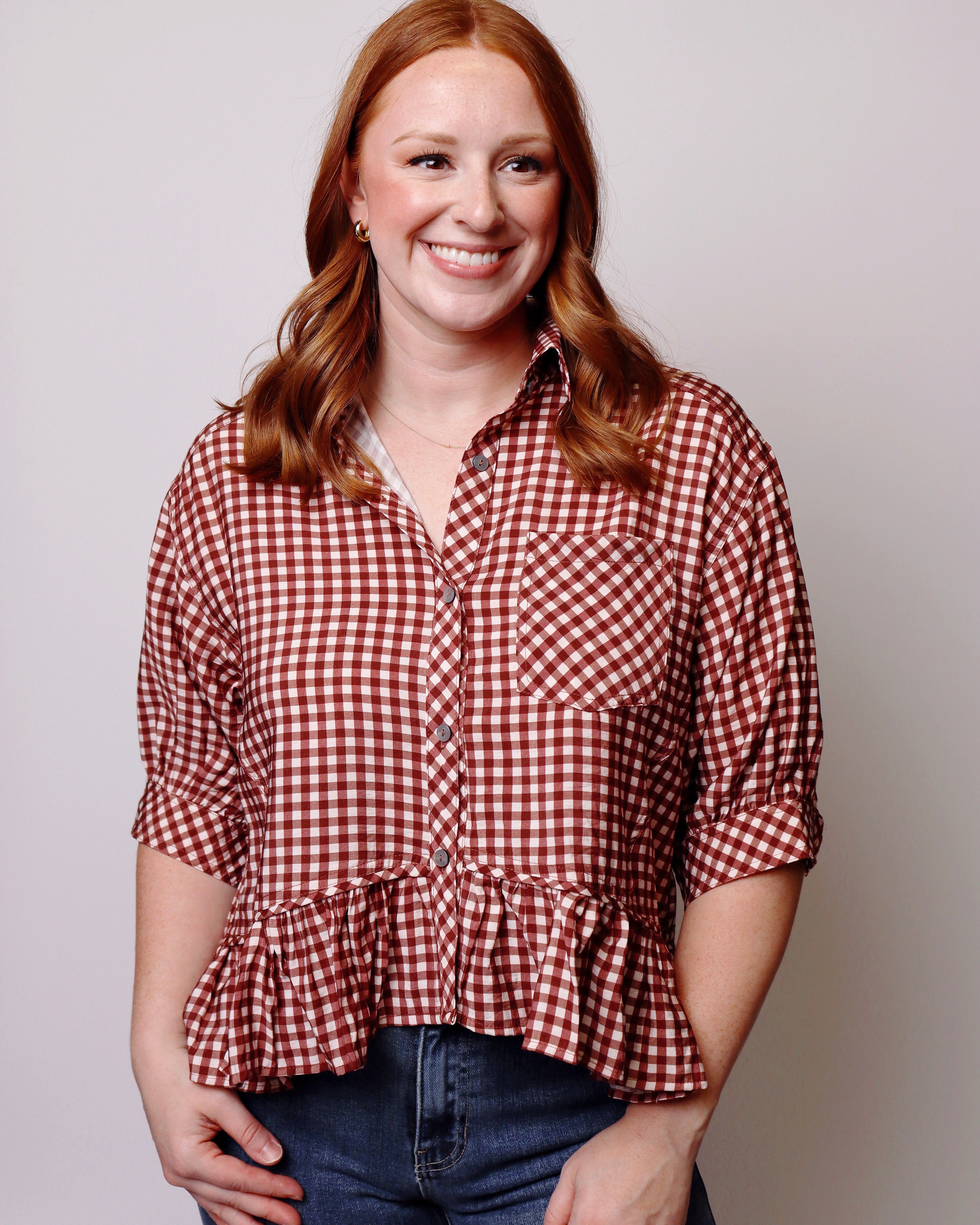A smiling woman with red hair wearing a red and white gingham check short sleeve blouse with a ruffled hem, paired with blue jeans, against a plain light gray background.