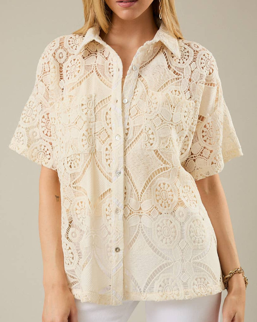 Woman wearing a beige lace blouse with white pants against a neutral background