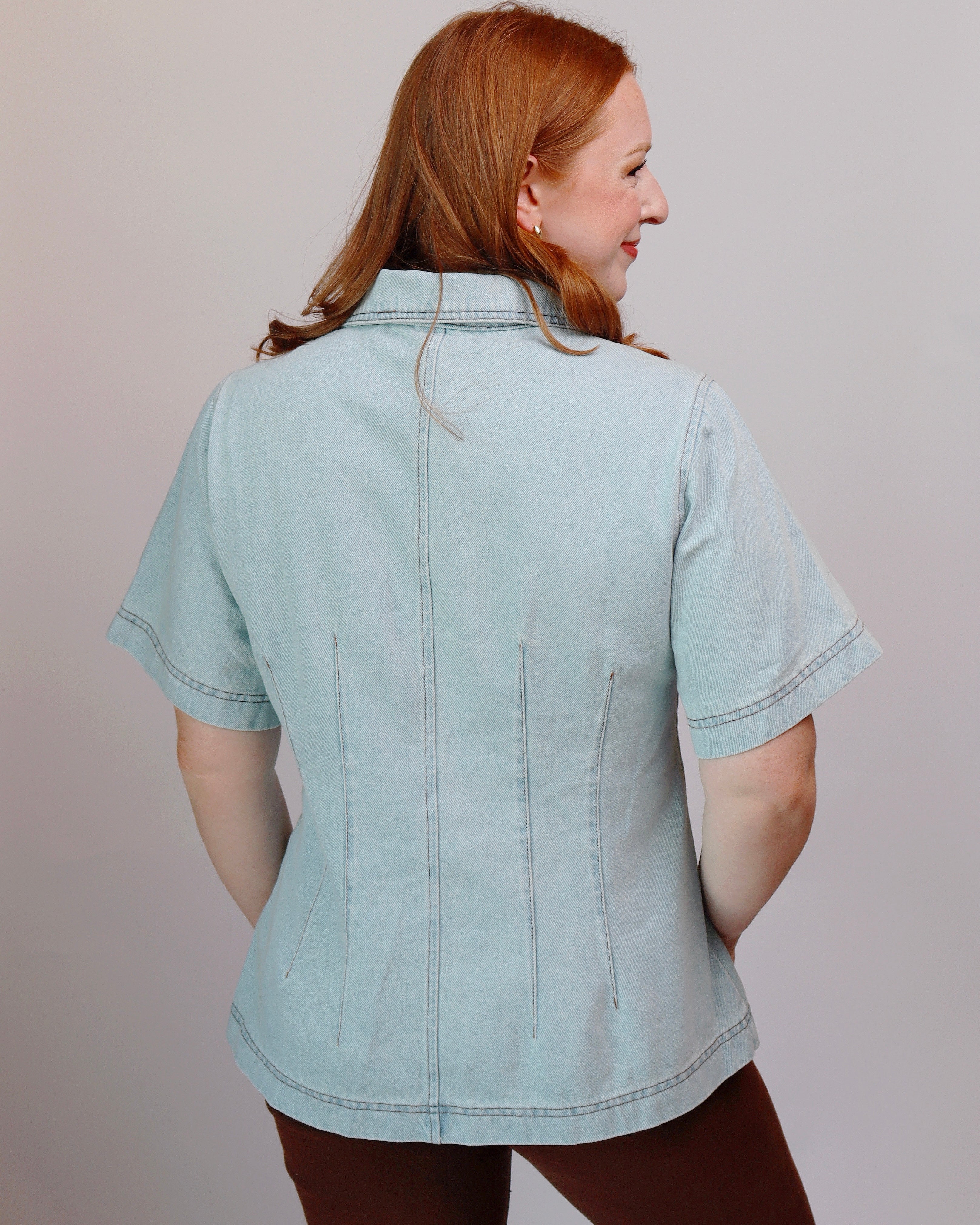 A woman with red hair is shown from the back wearing a light blue denim short-sleeve shirt with vertical seam detailing on the back, paired with brown pants, against a plain light gray background.