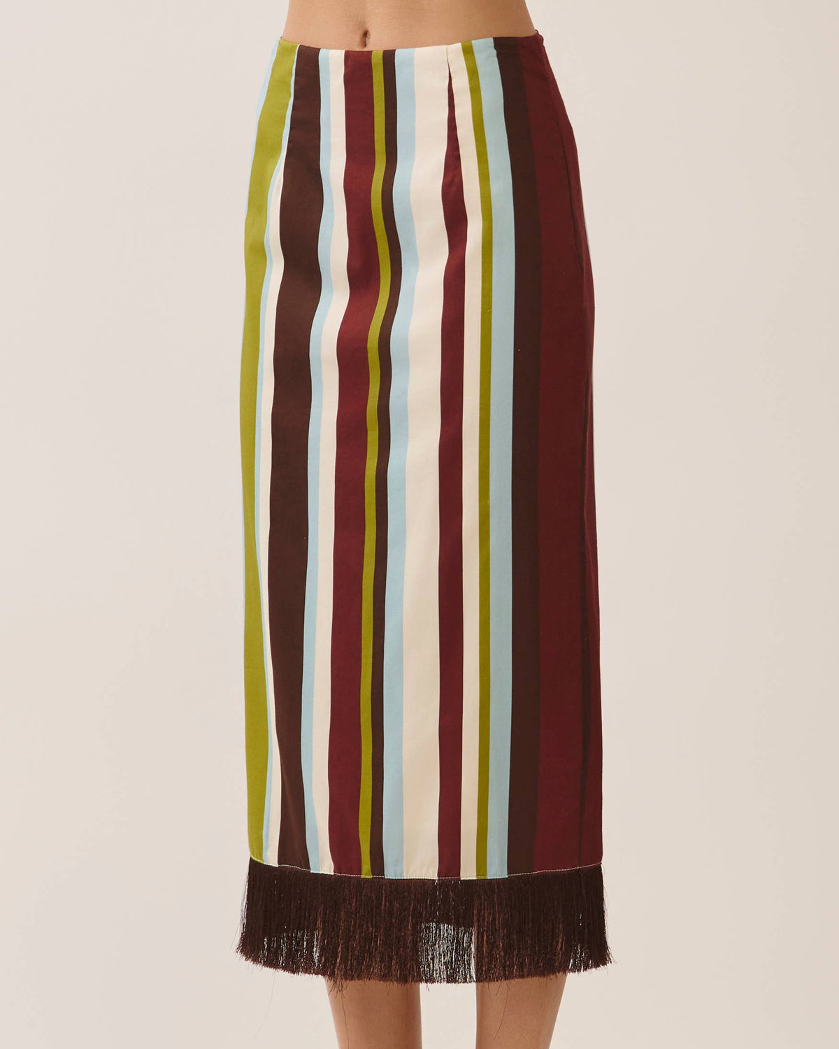 A model is wearing a midi skirt with vertical stripes in burgundy, olive green, cream, and light blue, finished with a dark brown fringe hem. Against a neutral background.