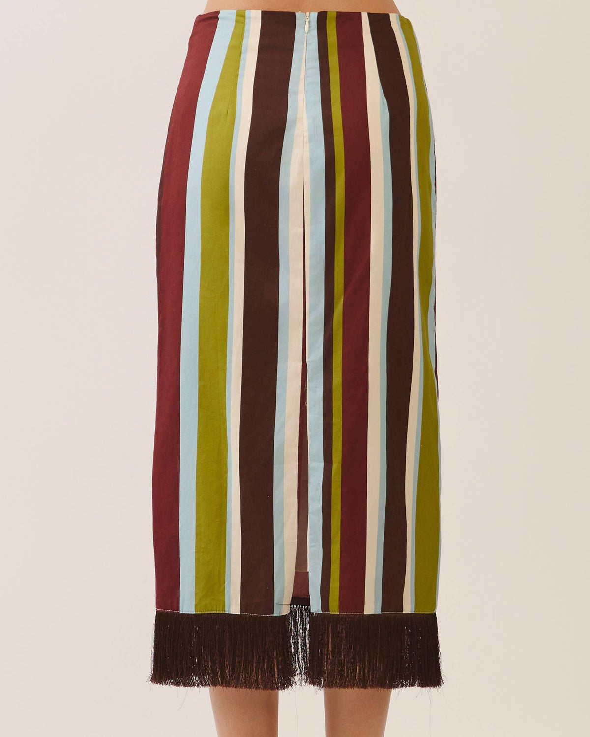 A model is wearing a midi skirt with vertical stripes in burgundy, olive green, cream, and light blue, finished with a dark brown fringe hem. Against a neutral background.