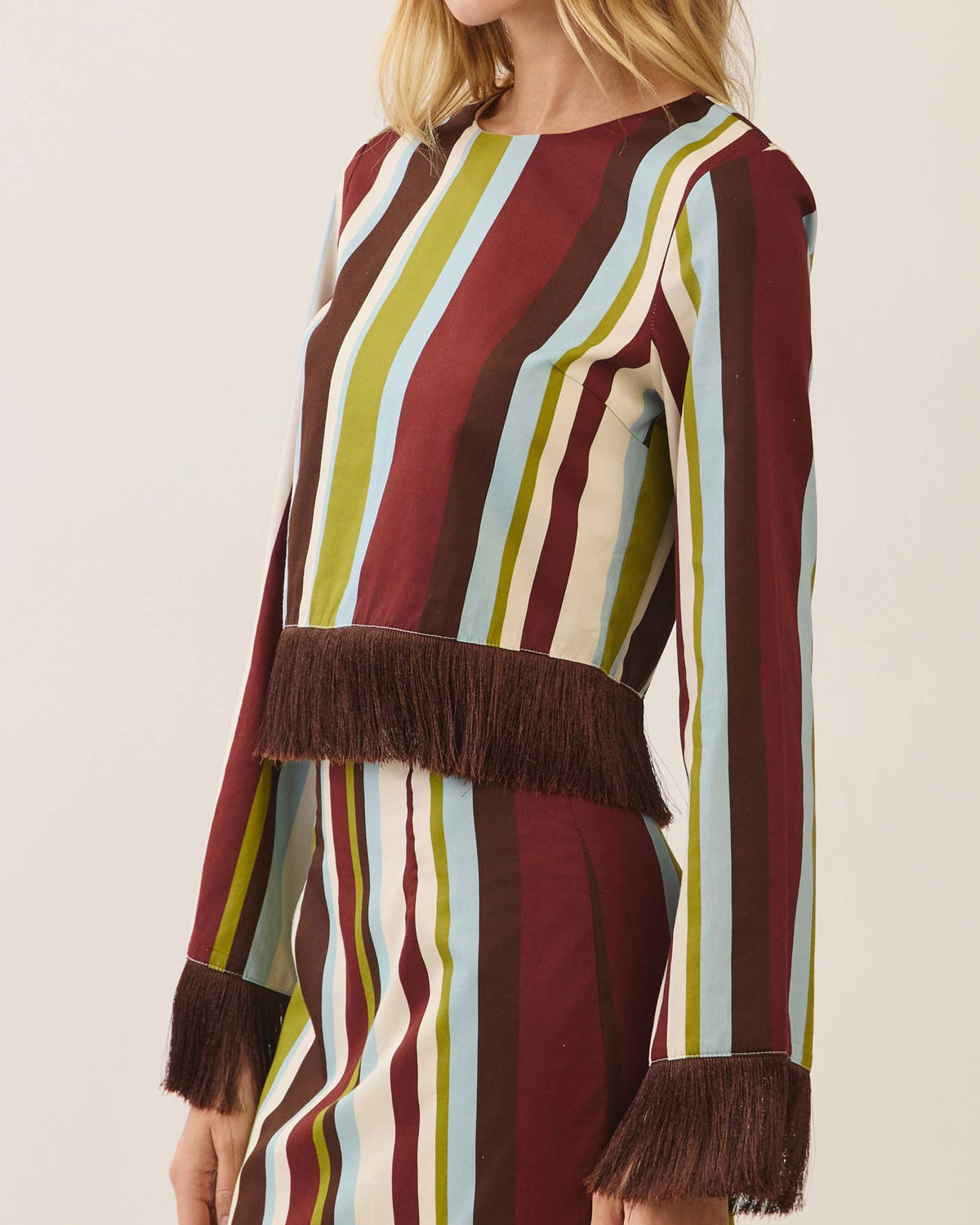 Model wearing a vertical striped long sleeve blouse with fringe at the cuff and hem on a neutral background.