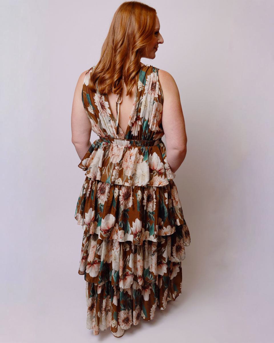 A woman with long red hair is shown from the back wearing a sleeveless, tiered maxi dress with a brown, beige, and green floral pattern against a plain light gray background.