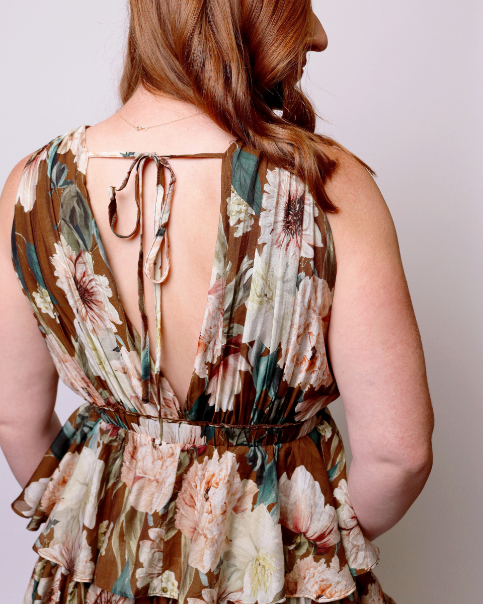 A woman shown from the back wearing a sleeveless floral dress with a tie-back neckline and a gathered waist, photographed against a plain light background.