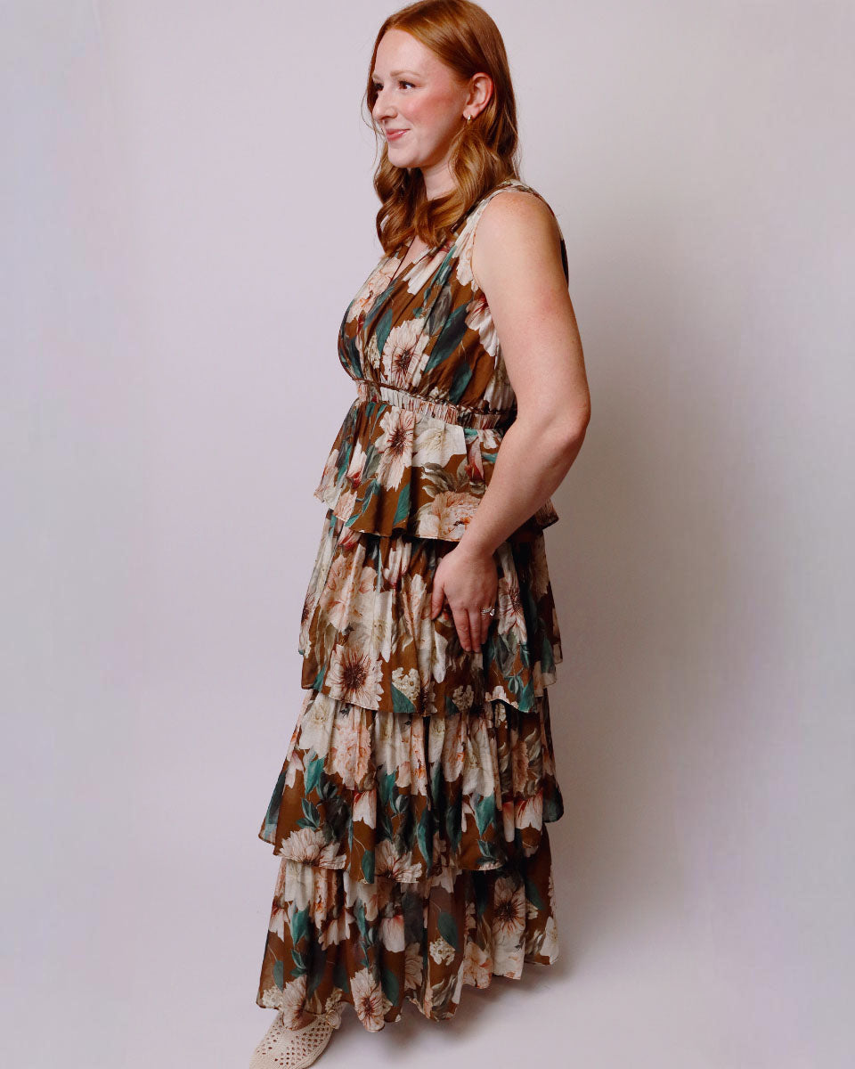 A woman modeling a sleeveless, tiered maxi dress with a brown, beige, and teal floral pattern, standing against a plain light gray studio background.
