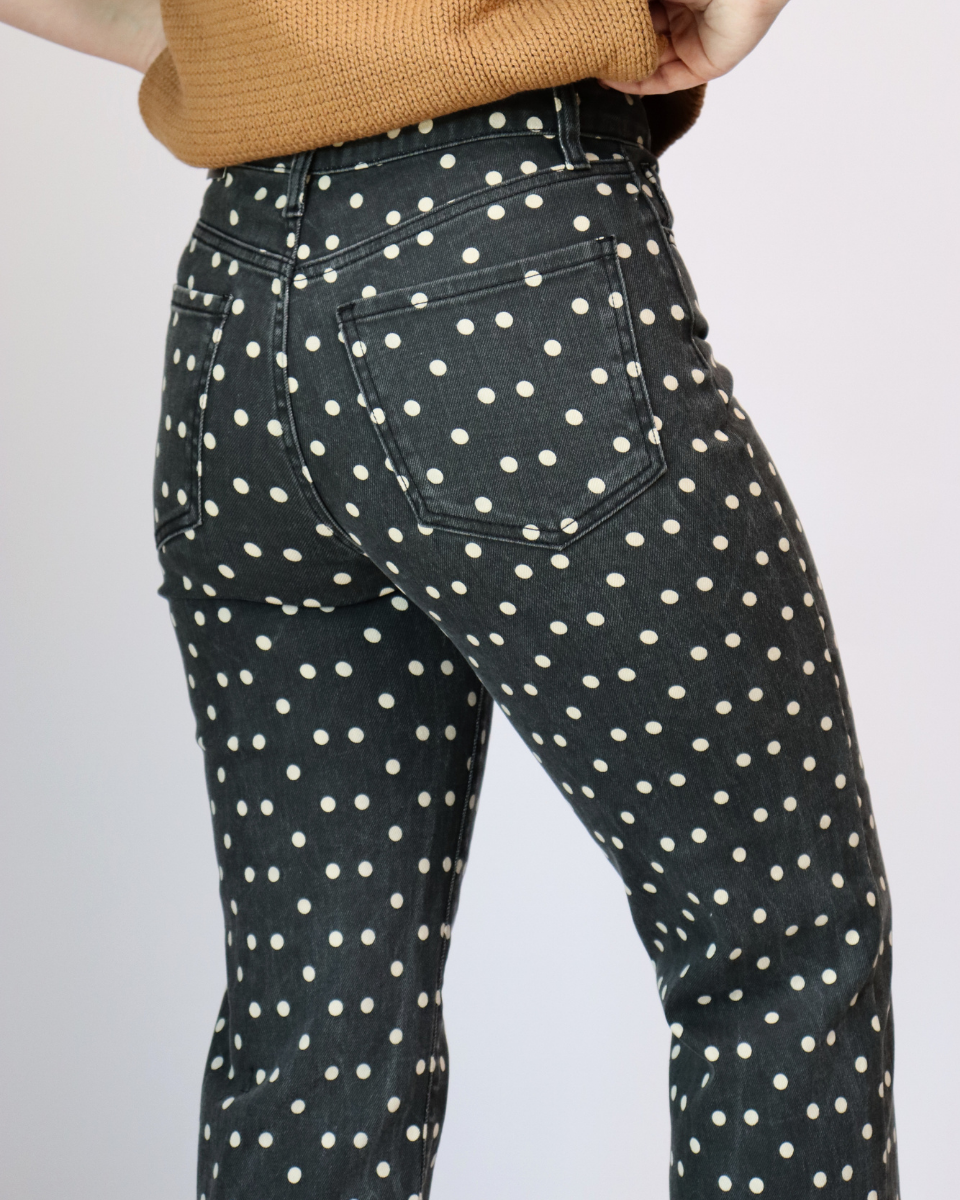 Close-up of a person wearing dark charcoal gray jeans with an all-over white polka dot pattern, paired with a tan ribbed sweater, shown from the back against a light gray studio background.