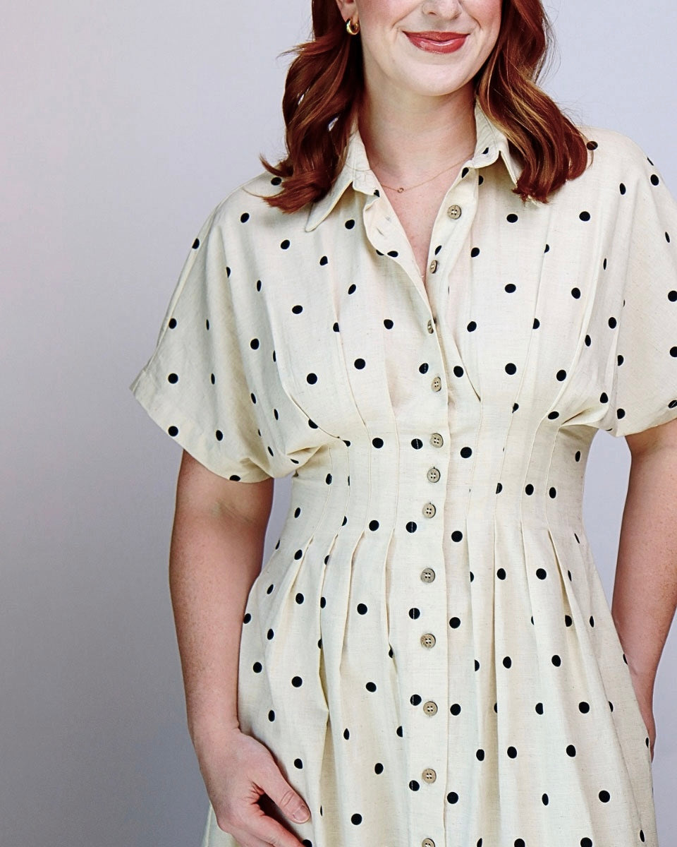 A woman wearing a cream-colored short-sleeve button-up dress with black polka dots, shown from the chest down against a plain light gray background.