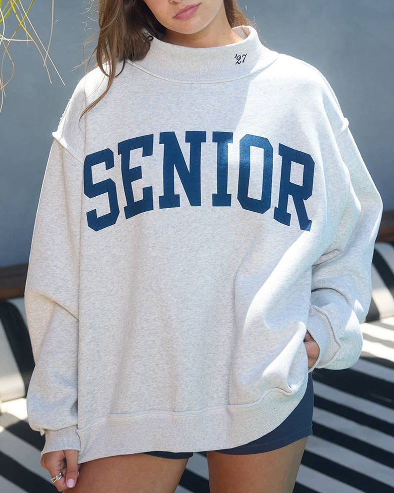 Person wearing a 'SENIOR' sweatshirt standing on a striped surface with an outdoor background.
