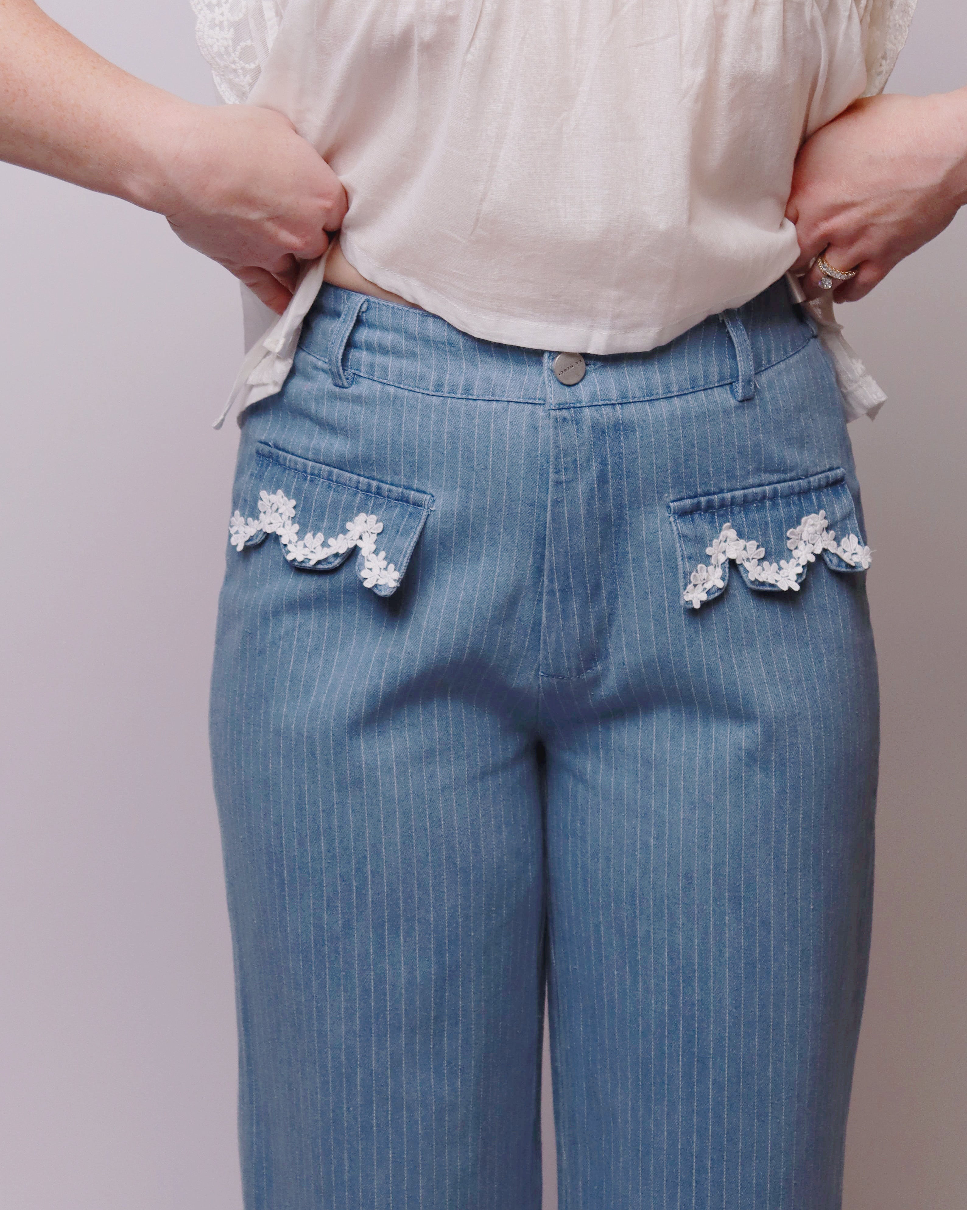 Blue pinstripe jeans with decorative pockets worn by a person on a plain background