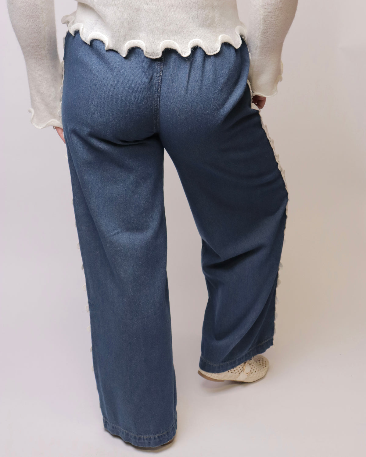 A person shown from the back wearing wide-leg blue jeans with a white ruffled hem sweater and beige shoes, photographed against a plain light studio background.