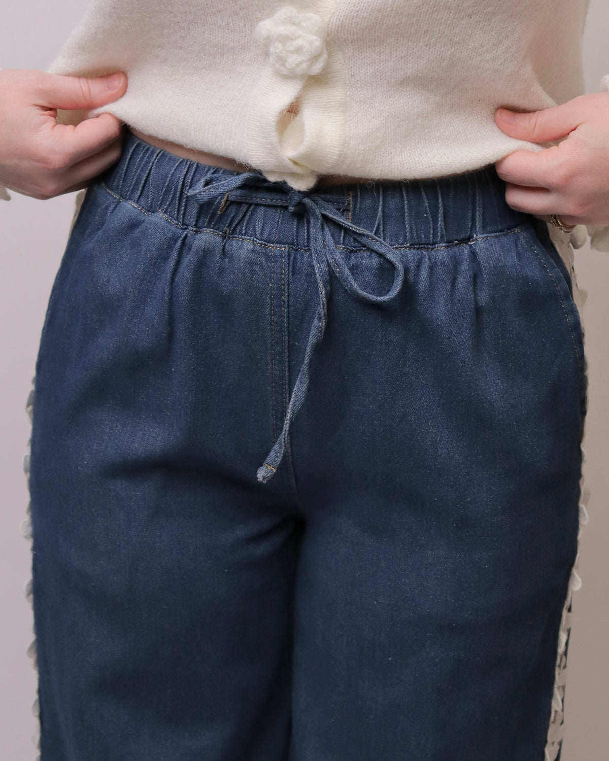 Close-up of a person wearing dark blue denim drawstring pants and a cream knit top, holding the waistband of the pants with both hands.