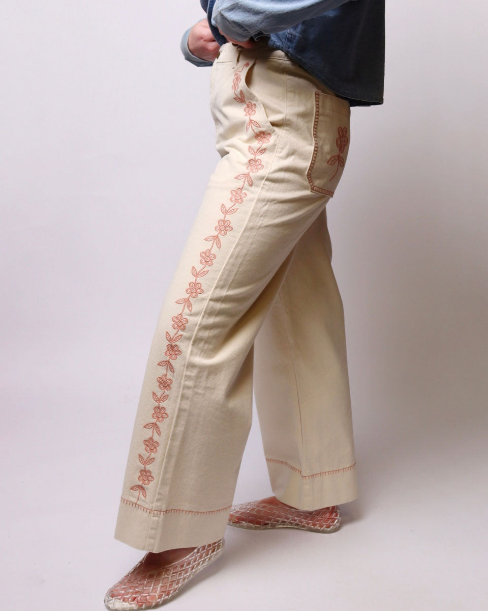 A person wearing cream wide-leg pants with pink floral embroidery down the outer sides, paired with a blue shirt and patterned slip-on shoes, photographed against a plain light gray studio background.
