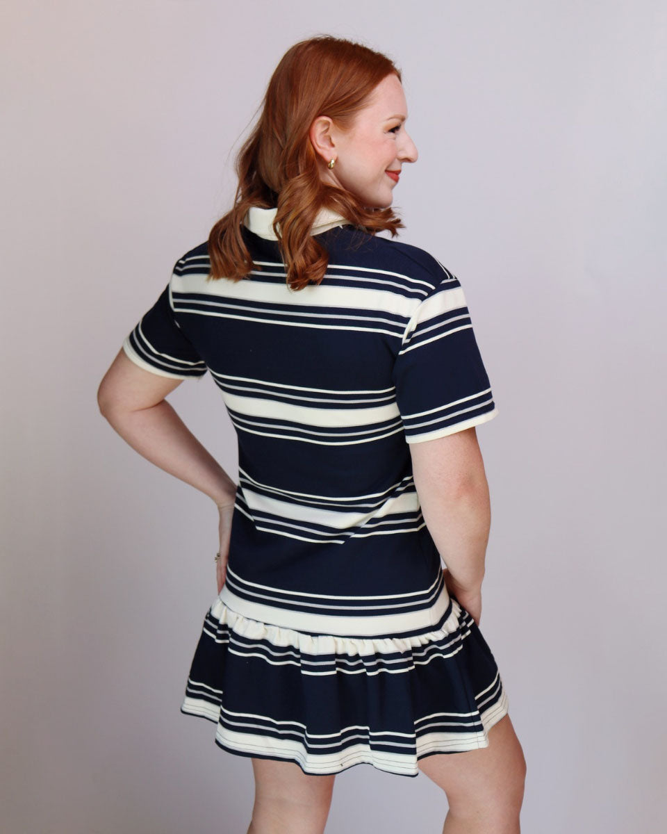 A woman with red hair is shown from the back wearing a short-sleeve ruffled polo dress with horizontal navy blue and cream stripes, set against a plain light gray studio background.