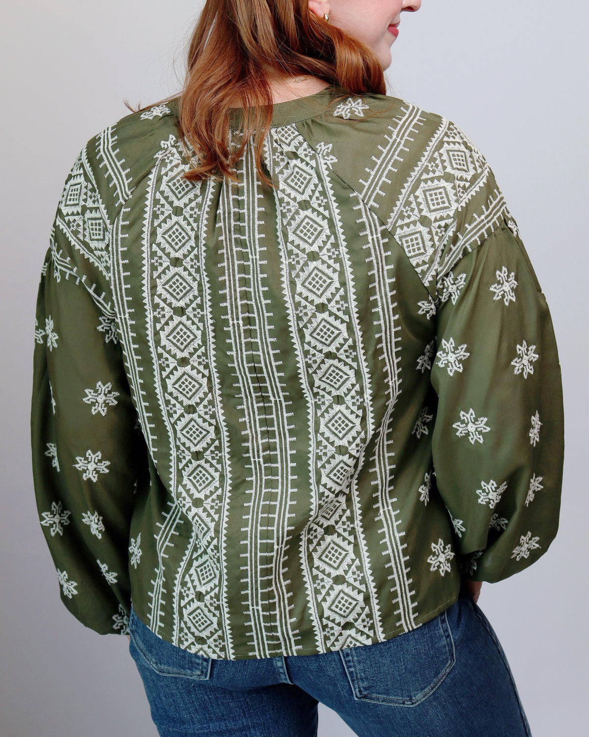 A woman wearing an olive green long-sleeve blouse with white geometric and snowflake patterns, shown from the back against a plain light gray background.