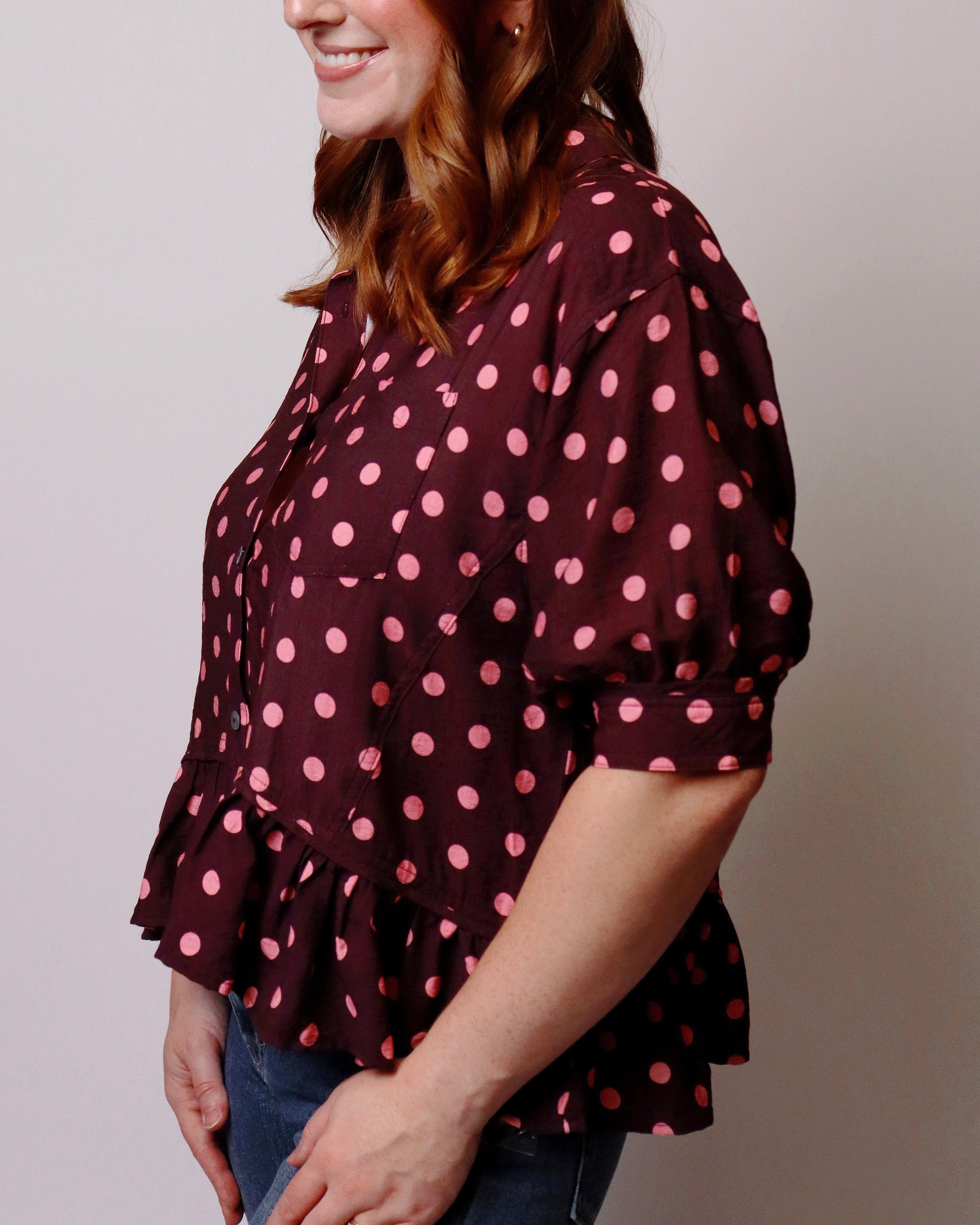 A woman wearing a dark burgundy blouse with pink polka dots, paired with blue jeans, shown from a side profile against a plain light gray background.