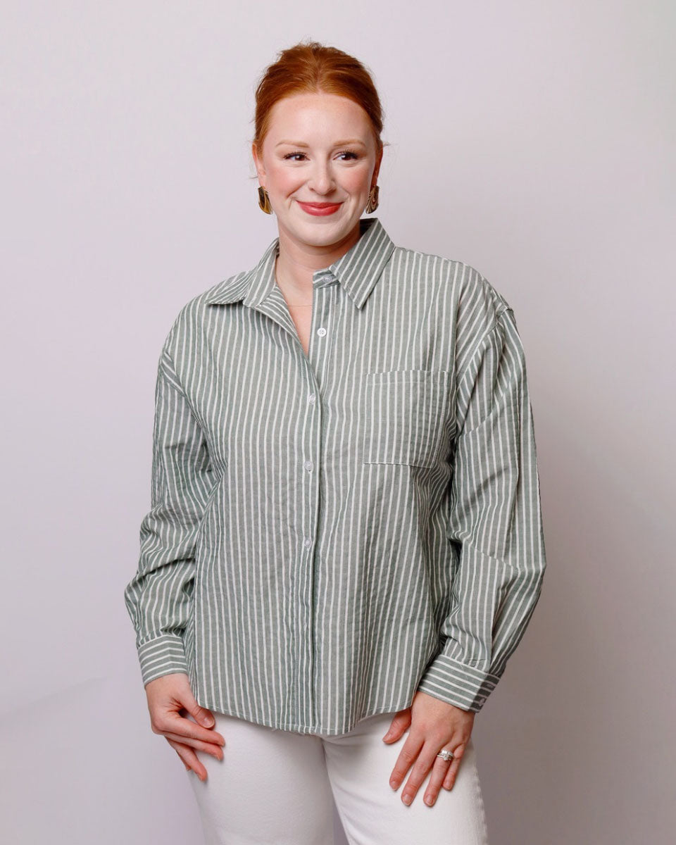 A woman with red hair wearing a sage green and white vertically striped button-down shirt and white pants, standing against a light gray studio background.