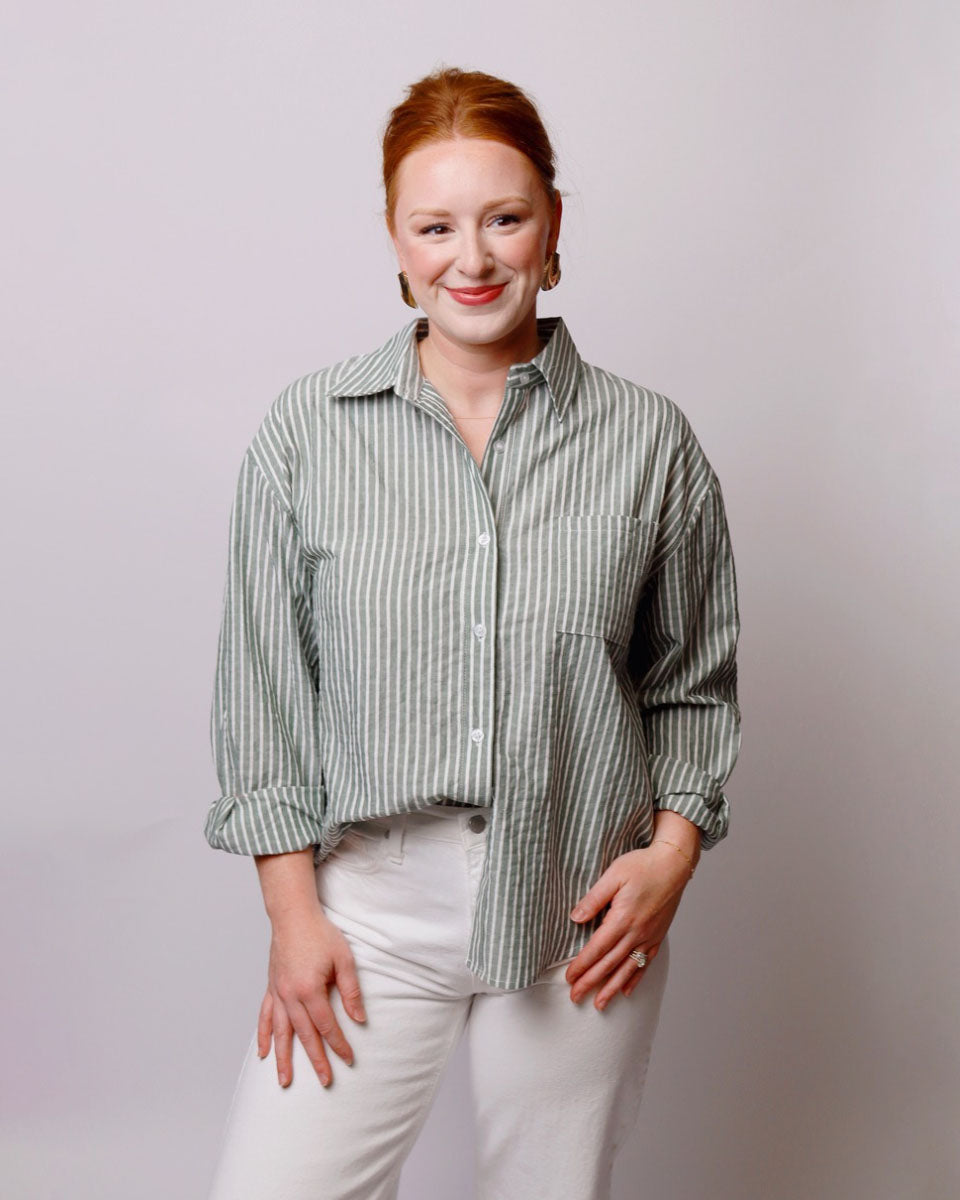 A woman with red hair tied back, wearing a sage green and white vertically striped button-down shirt with white pants, standing against a light gray studio background.
