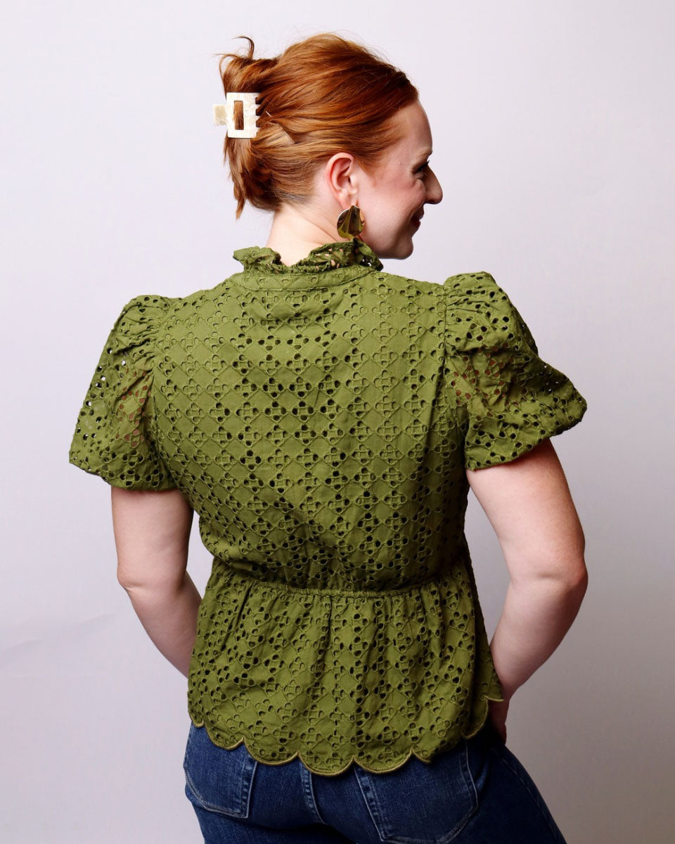A woman with red hair tied up in a bun is shown from the back, wearing an olive green eyelet lace blouse with puff sleeves and a ruffled high neck, paired with blue jeans against a plain light gray background.