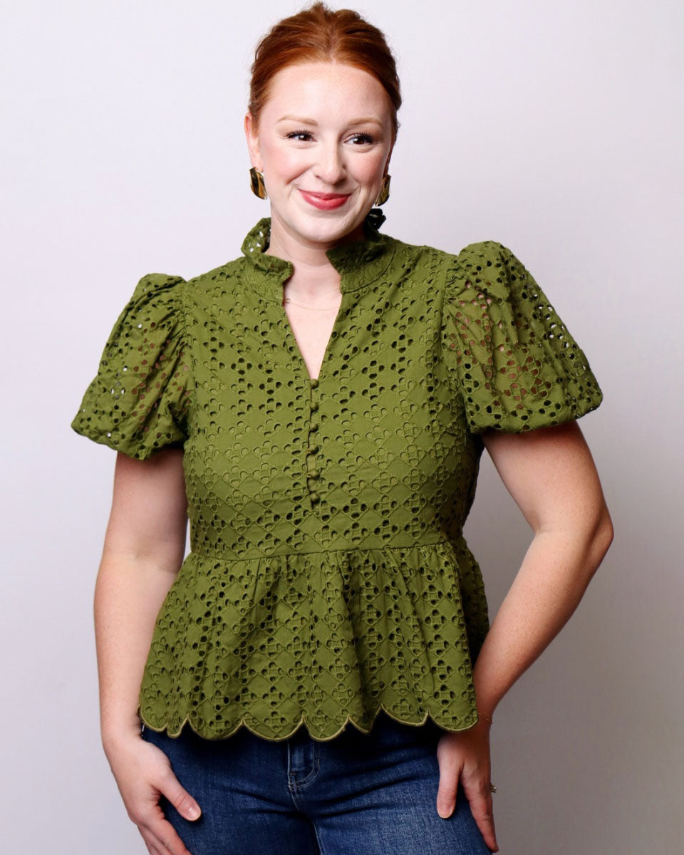 A woman with red hair wearing an olive green eyelet lace blouse with puff sleeves and a scalloped peplum hem, paired with blue jeans, against a light gray studio background.
