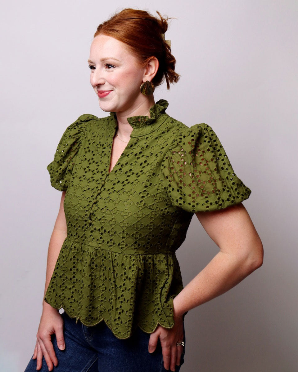 A woman with red hair in a bun wearing an olive green eyelet lace blouse with puff sleeves and a scalloped peplum hem, paired with blue jeans, posing against a light gray studio background.