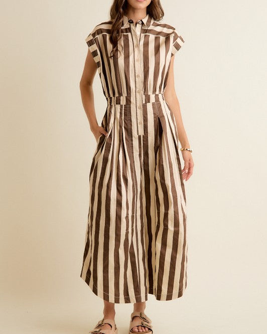 A woman modeling a brown and beige vertically striped jumpsuit with a button-up collar and sleeveless cap sleeves, paired with brown platform sandals, against a solid cream background.