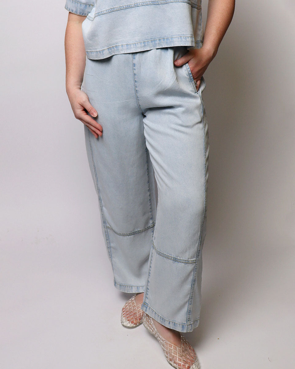 A person wearing light blue wide-leg denim pants with contrast stitching, paired with a matching light blue top and beige lace-up flats, shown against a plain light gray studio background.