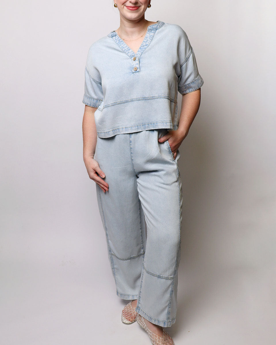 A woman modeling a light blue denim two-piece set consisting of a short-sleeve cropped top with decorative trim and buttons, and matching wide-leg jeans, against a plain light gray studio background.