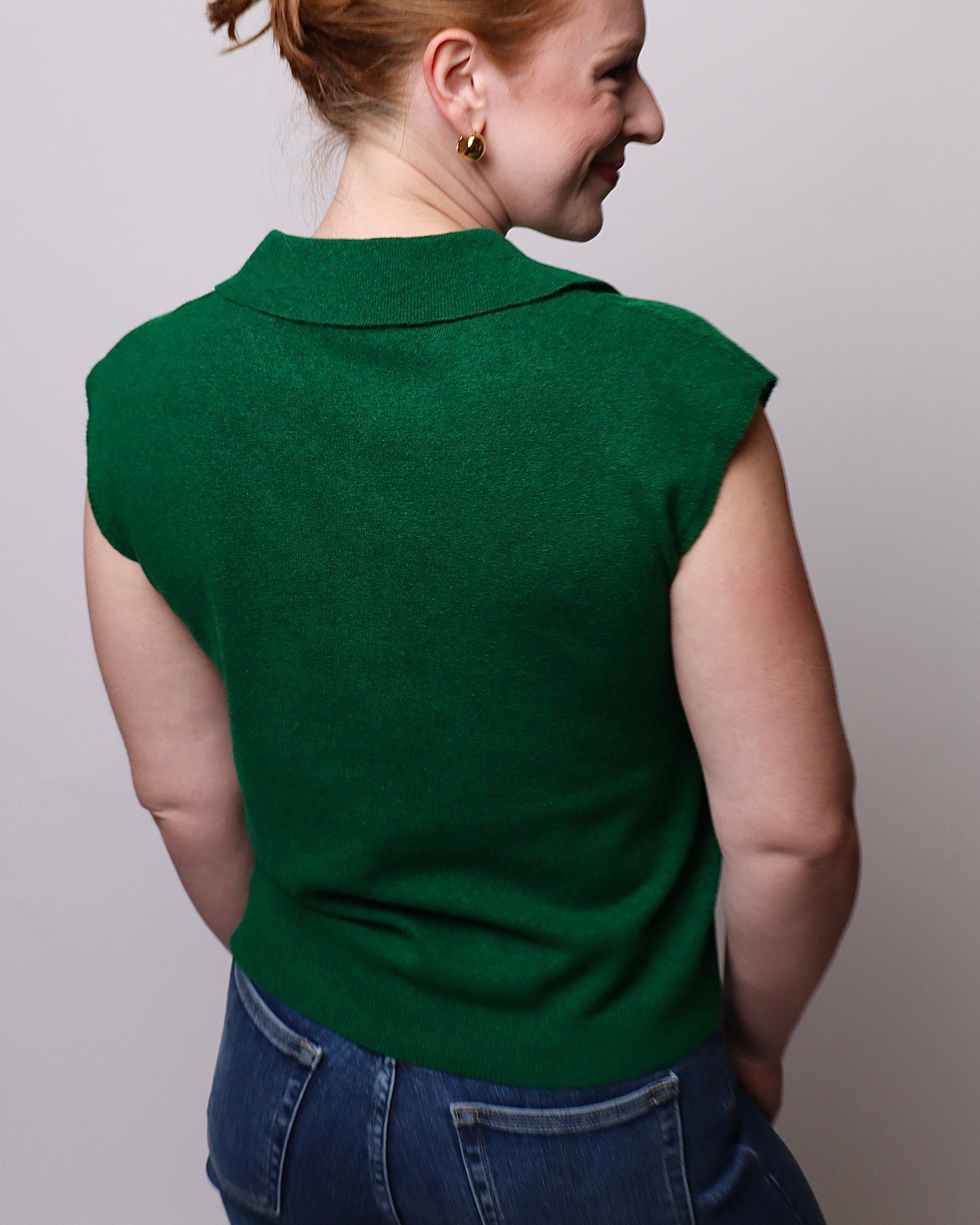 A woman shown from the back wearing a dark green sleeveless knit top with a collar, paired with blue jeans, against a plain light gray background.