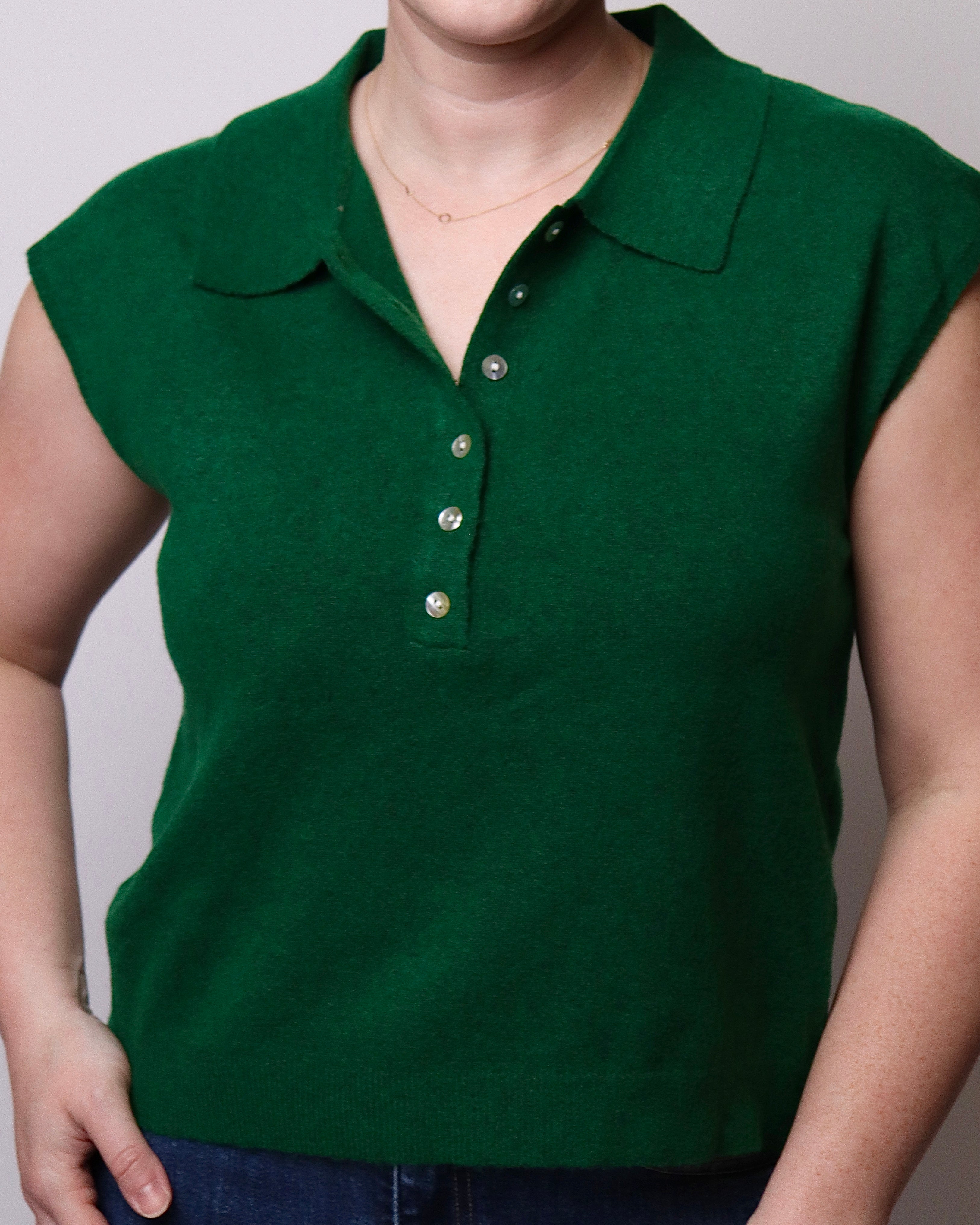 A close-up of a person wearing a dark green sleeveless polo shirt with a collar and button placket, paired with blue jeans against a plain light gray background.