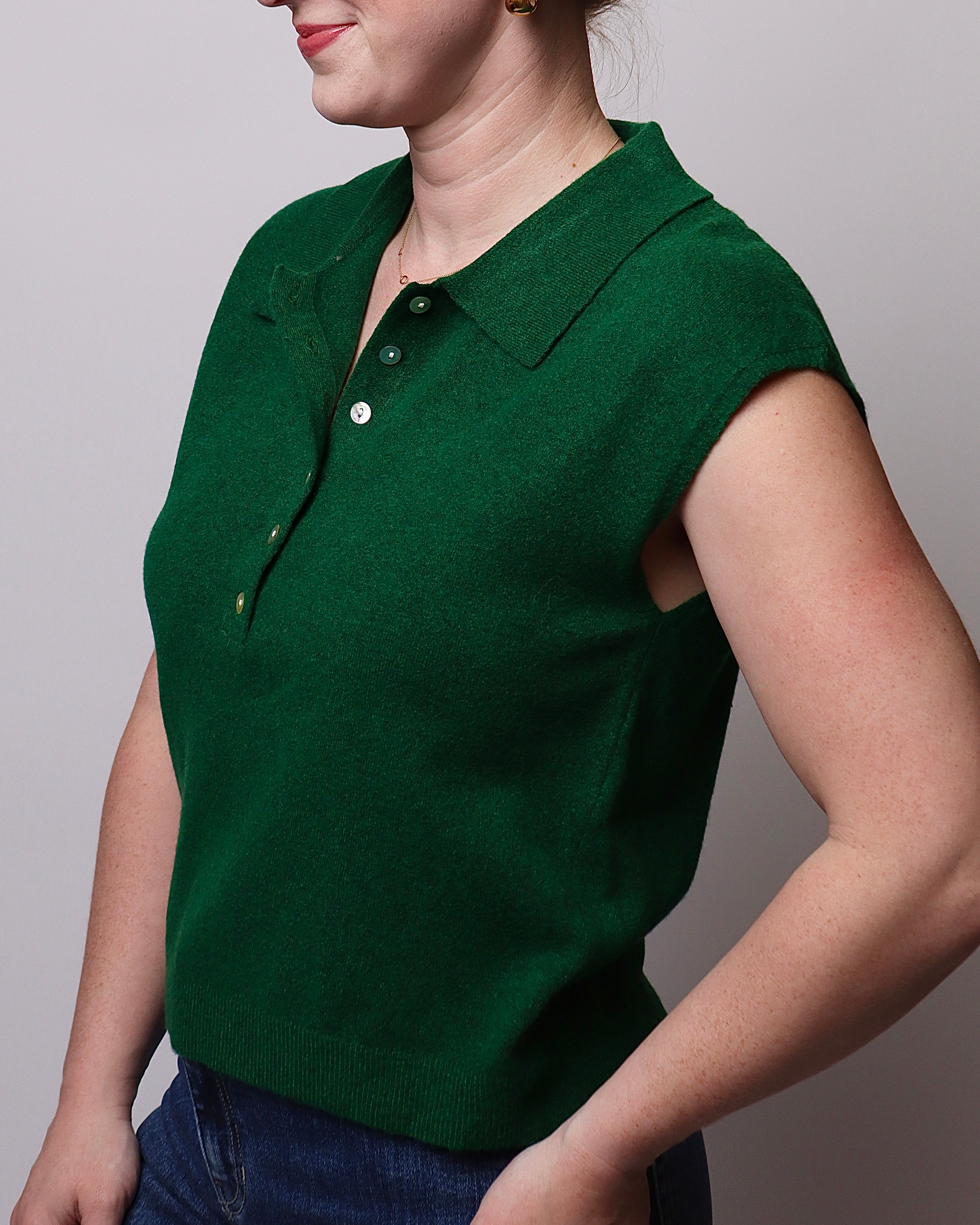 A woman wearing a dark green sleeveless knit polo sweater with a short button placket, paired with blue jeans, shown against a plain light gray background.