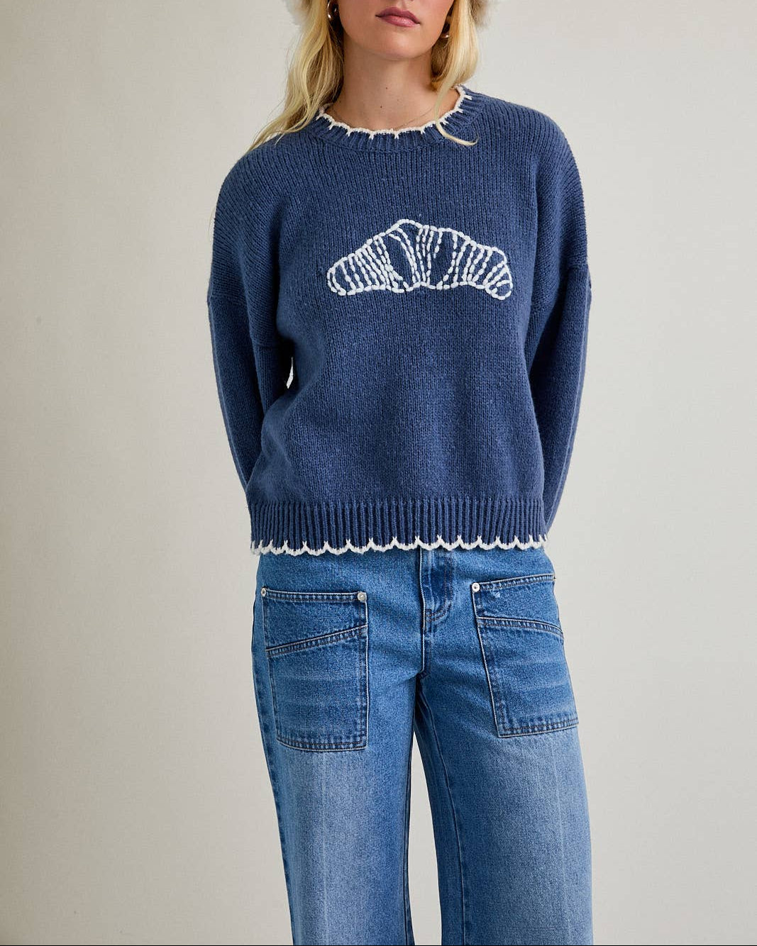 Woman wearing a blue sweater with a white design, blue jeans, and a white fur hat on a plain background