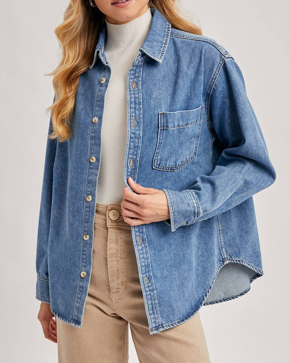 Woman wearing a blue denim shirt over a white turtleneck and beige pants on a light gray background