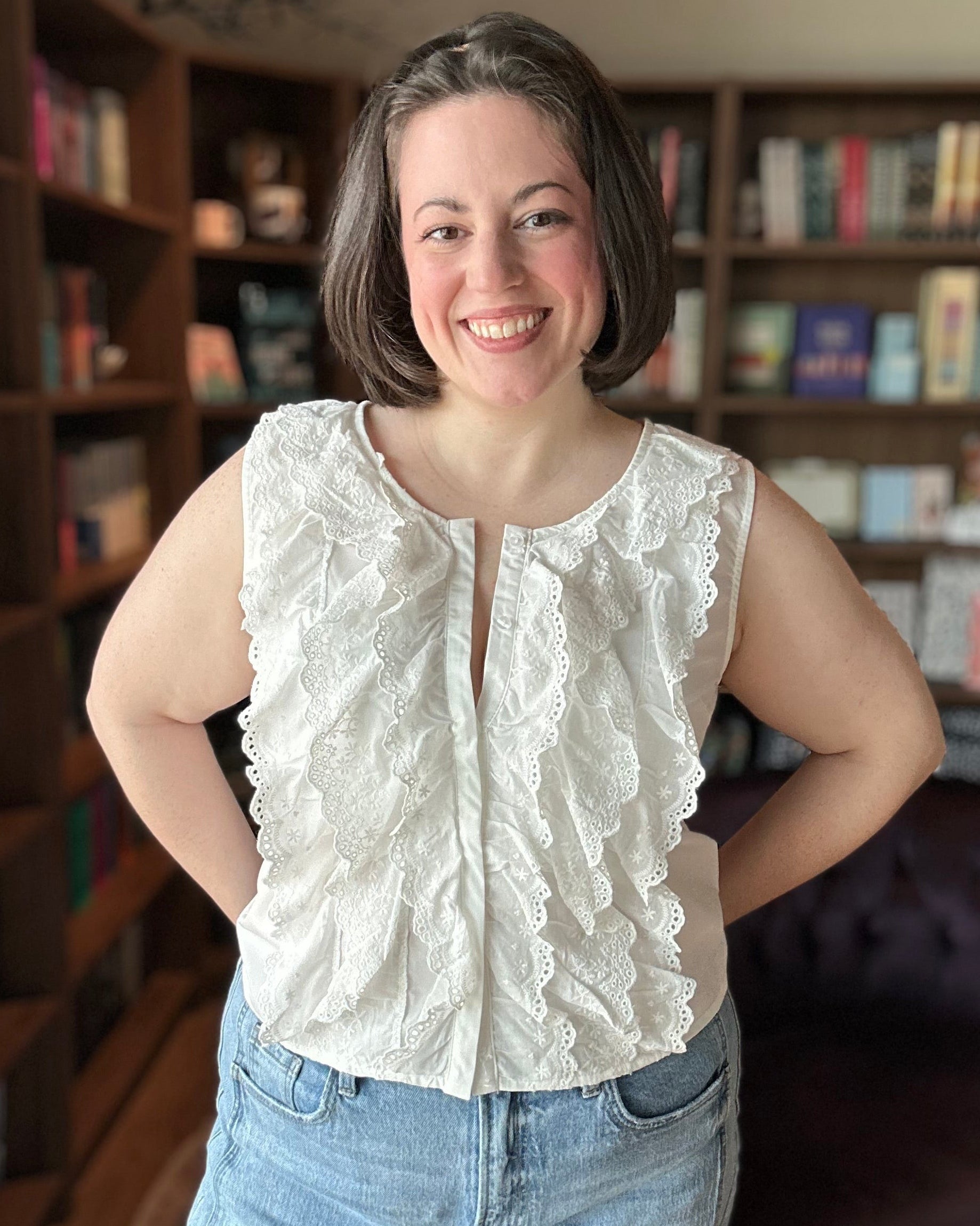 Woman wearing a white sleeveless top and blue jeans standing in a room with bookshelves.