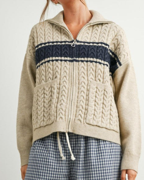 Woman wearing a beige cable knit sweater with navy stripe and plaid pants on a white background