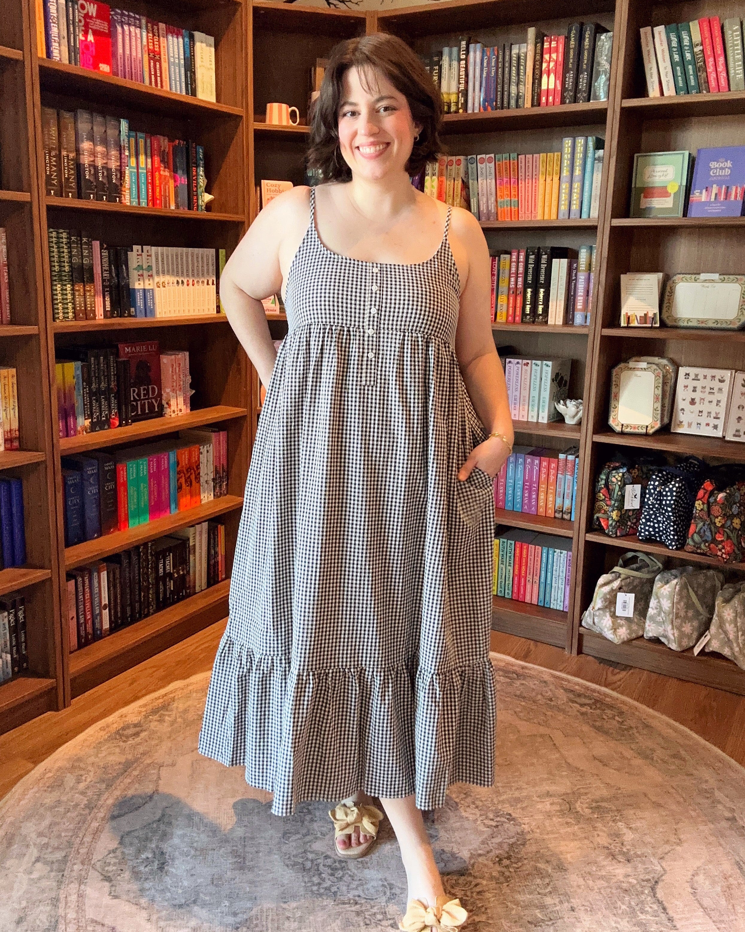Woman in a checkered dress standing in front of a bookshelf filled with books and decorative items.