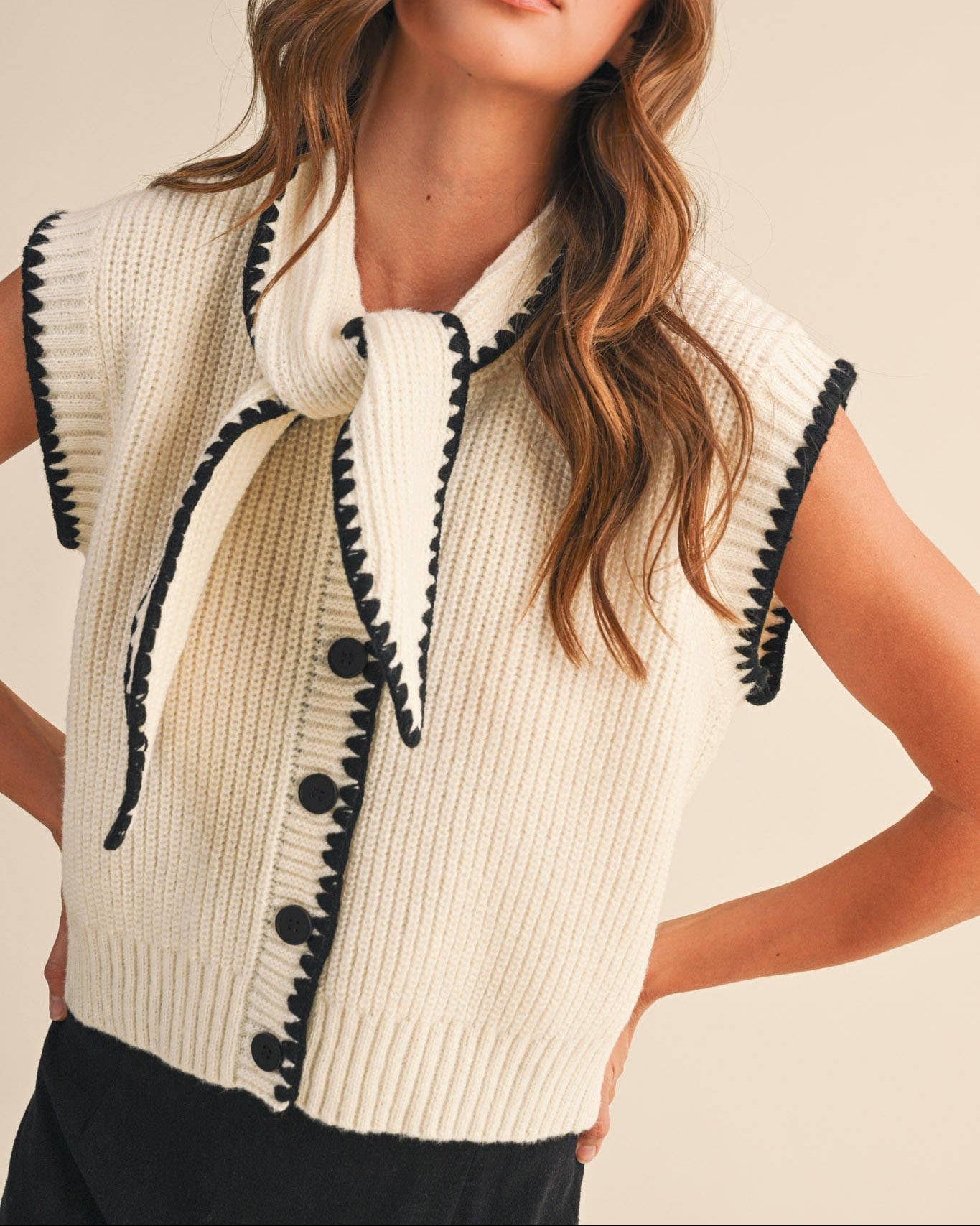 Woman wearing a cream-colored sleeveless sweater with black trim against a plain background
