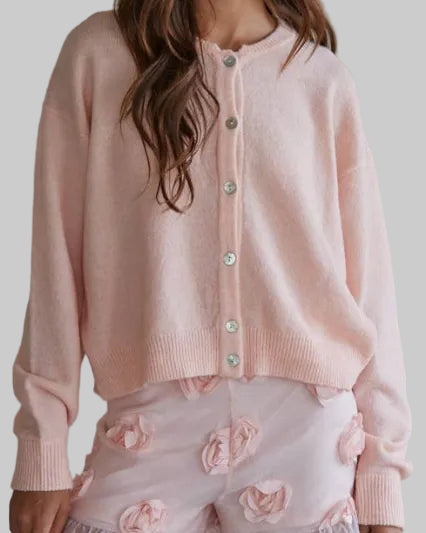 Woman wearing a pink cardigan and white shorts with floral details against a plain background