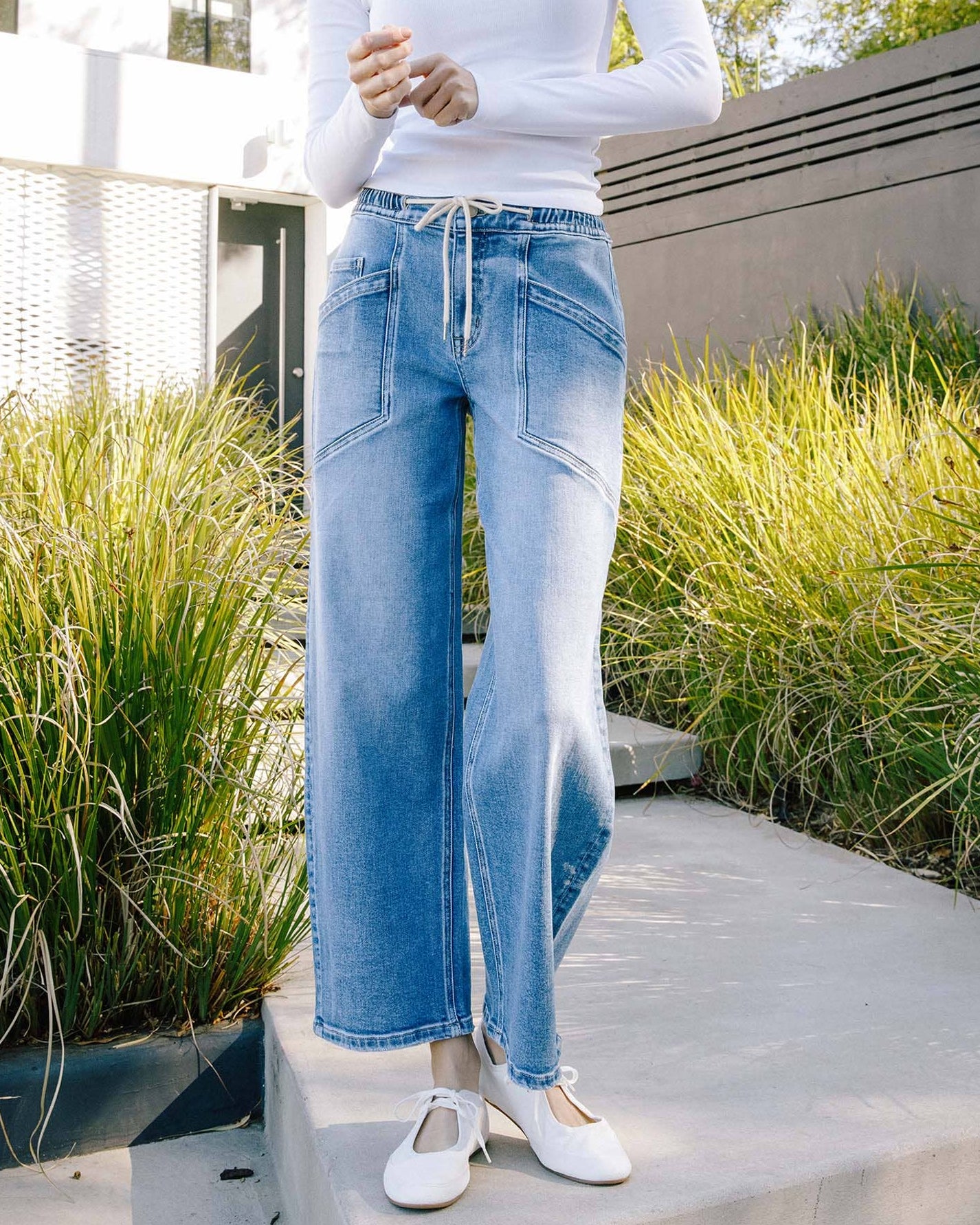 Woman wearing a white long-sleeve top and blue jeans standing outdoors with greenery in the background