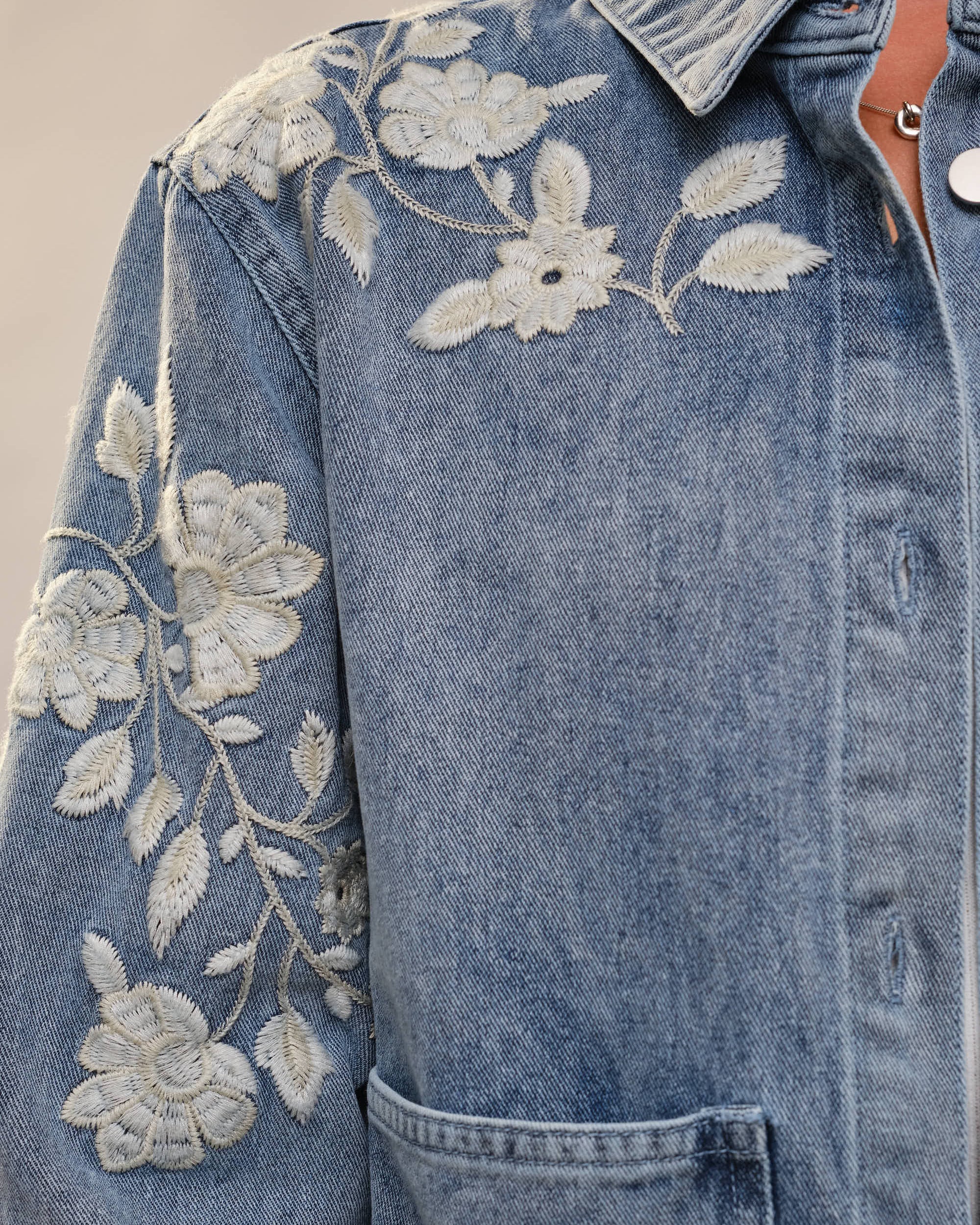 Blue denim jacket with white floral embroidery on a neutral background