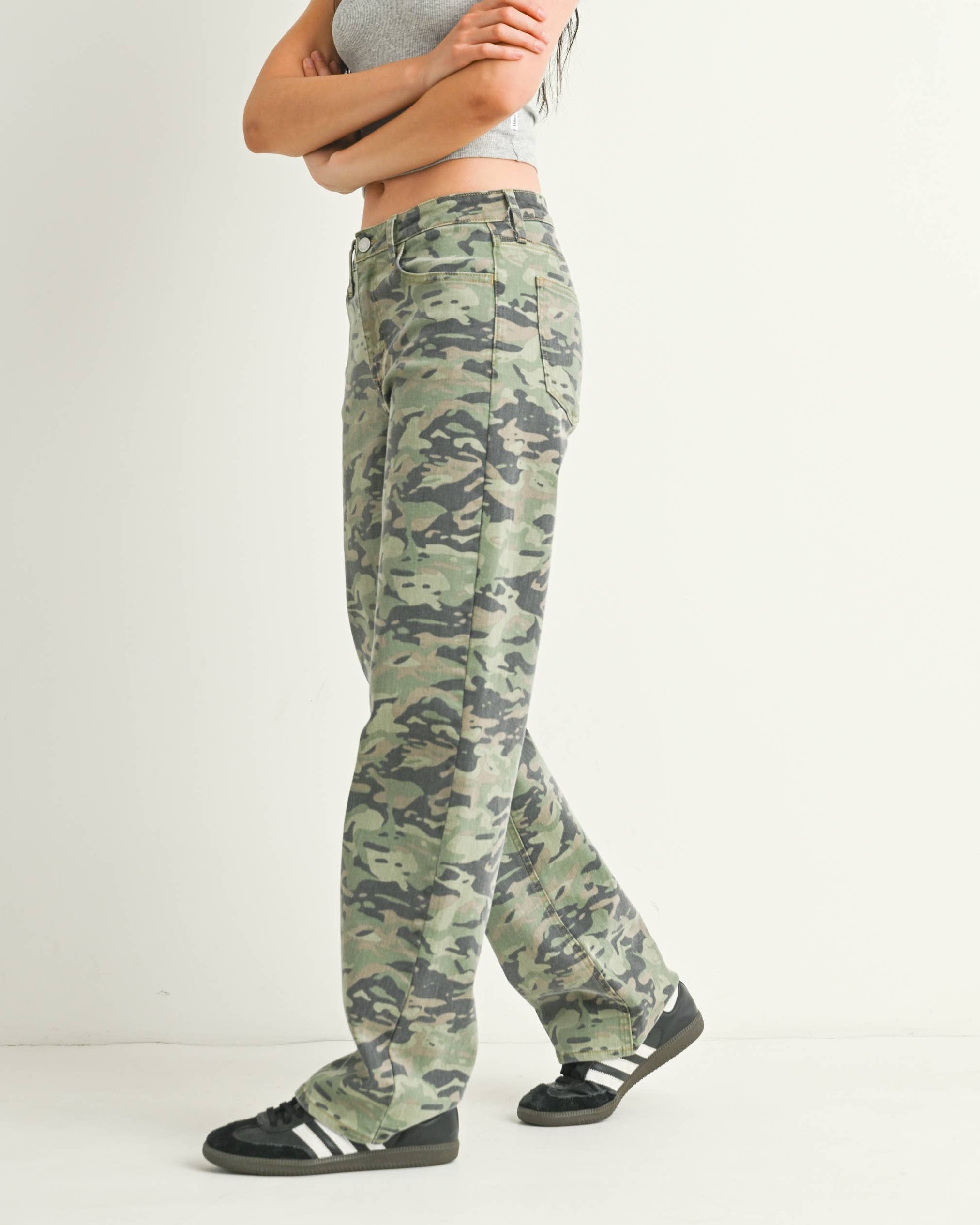 A woman modeling a pair of green camouflage cargo pants, paired with a gray tank top and black sneakers, standing against a plain white studio background.