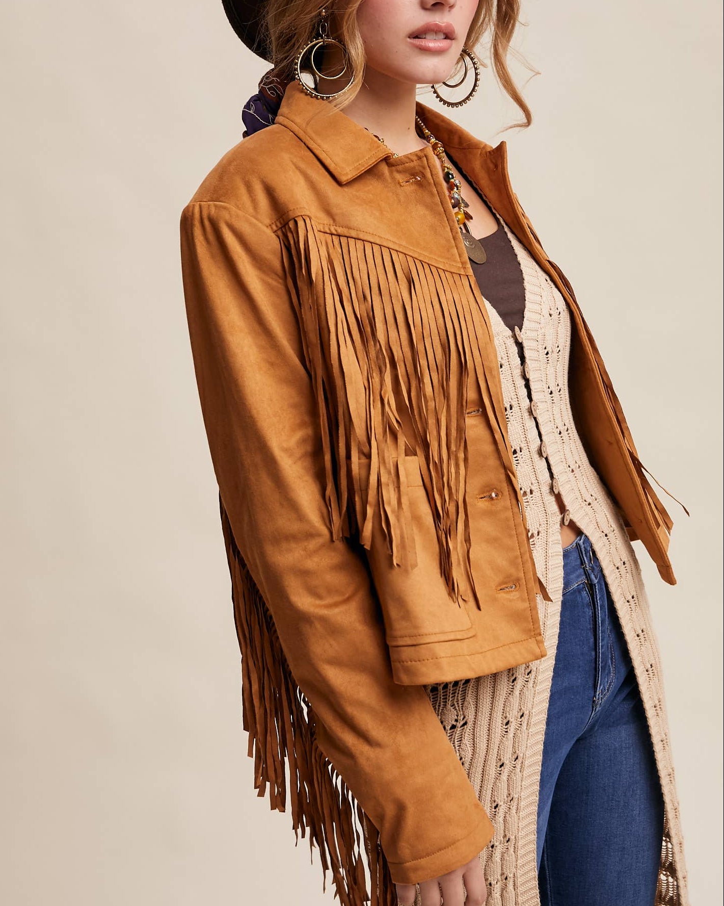 Woman wearing a brown fringed jacket, hat, and cardigan on a beige background